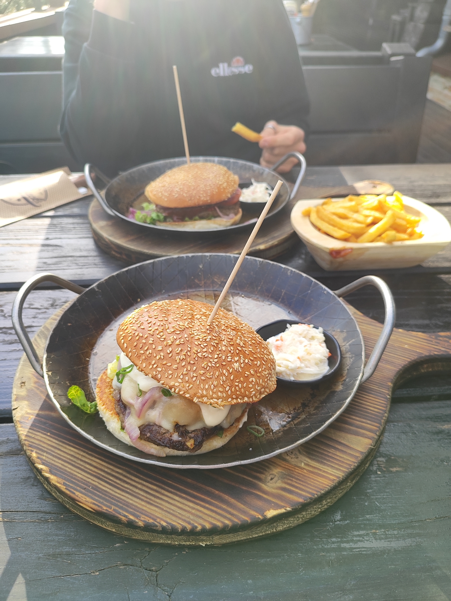 The image shows a wooden table setting with two burgers and a side of fries. Each burger is served in a metal pan placed on a wooden board. Both burgers have a sesame seed bun and are secured with a wooden skewer. A small black dish containing coleslaw accompanies the burger in the foreground. A separate wooden container holds a portion of fries. The person in the background is holding a single french fry. The location is the city of Unknown, Unknown.