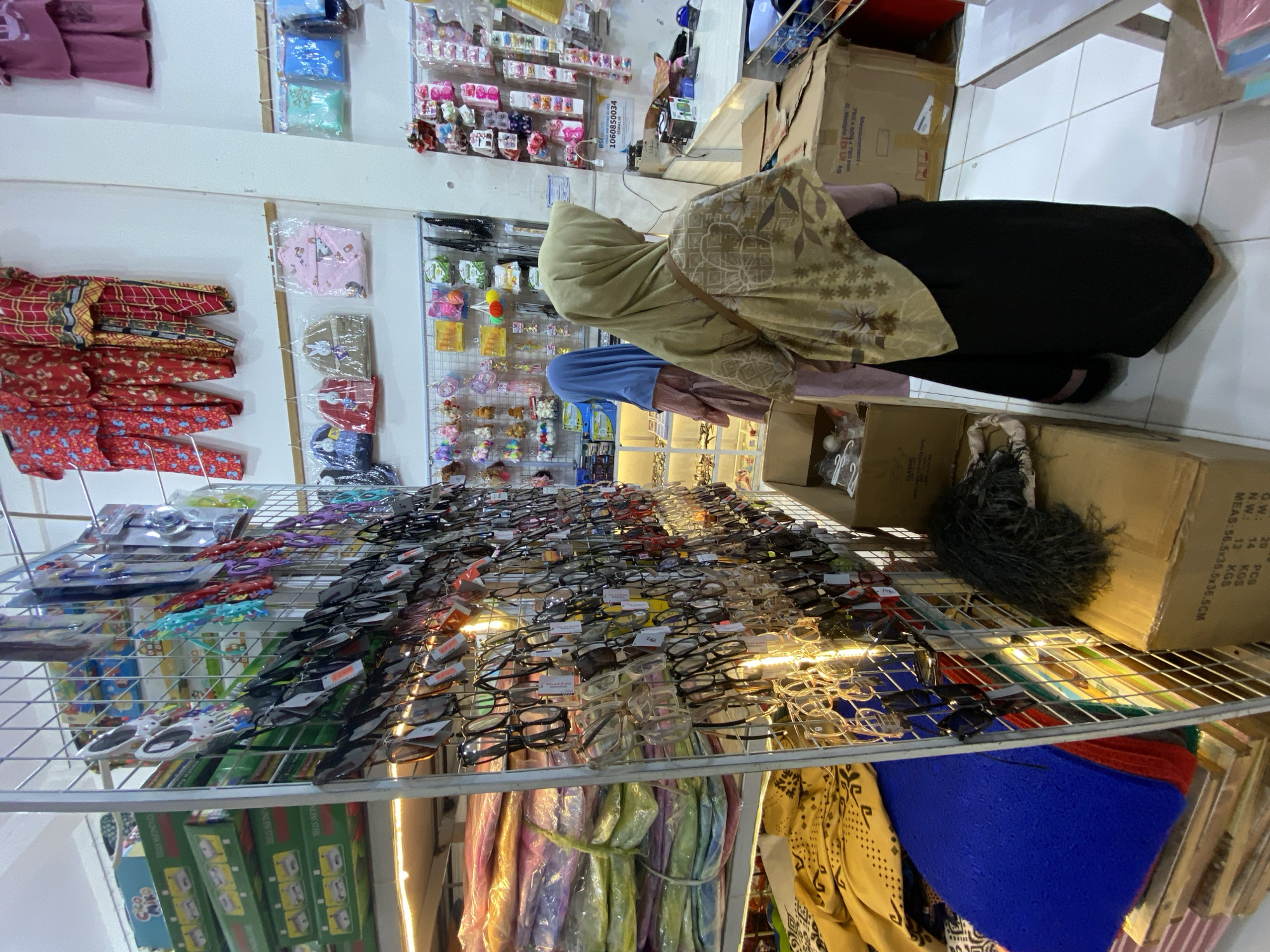 This is an eye-level, indoor shot of a store aisle filled with merchandise. A person, likely a woman based on the hijab and clothing, is seen from the back, wearing a light brown hijab and a black garment. She appears to be browsing or shopping. The aisle is lined with shelves displaying a variety of items. The shelves on the left hold colorful fabrics or clothing, possibly garments like sarongs or dresses, folded neatly. The shelves on the right are packed with rows of eyeglasses, with many pairs displayed on racks. Some of the eyeglasses have price tags attached. In the background, more shelves hold various small items, possibly accessories or toys, including what look like keychains with animal figures. Cardboard boxes are visible on the floor and on some of the shelves. One box has text that includes "Measurement 760 x 600 x 780 mm" and "GW: 20 PCS N.W.: 13 KGS MEAS: 56.5x35.5x36.5CM". The lighting suggests it is daytime or that the store is well-lit. The overall impression is of a busy retail environment, likely a small shop or market stall, possibly in Indonesia given the location context. The scene conveys a sense of everyday commerce.