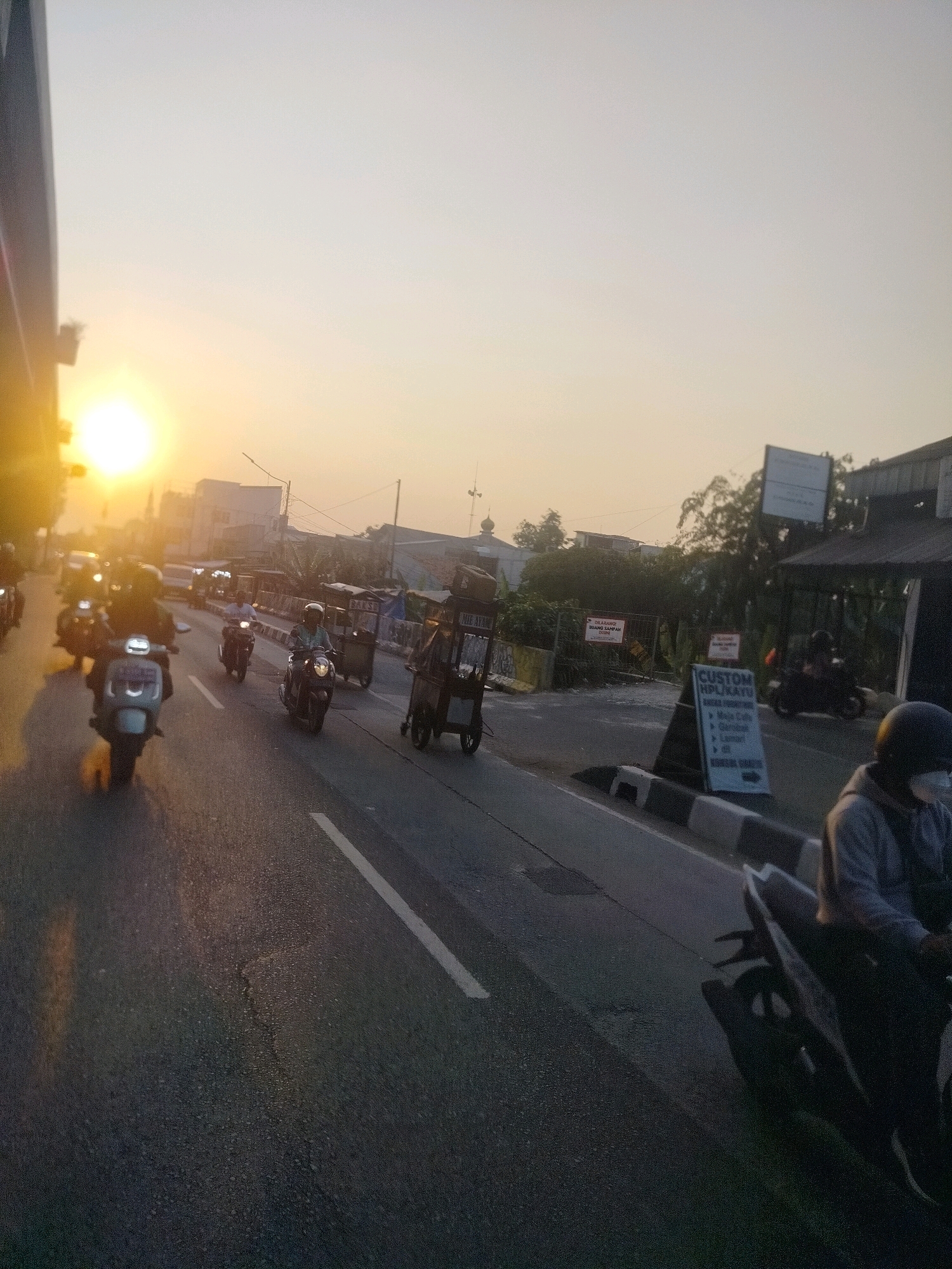 The image depicts a street scene in East Jakarta, Indonesia, during what appears to be sunrise or sunset, given the low angle of the sun and the warm, hazy light illuminating the sky. A busy road is visible, with multiple motorcycles navigating the traffic. The foreground features a person on a motorcycle, facing away from the camera and wearing a grey hoodie and a dark helmet with a mask. The road itself is asphalt with white lane markings.
On the right side of the road, there are structures that suggest commercial activity. A sign with "CUSTOM HPL/KAYU" is prominently displayed, indicating custom furniture or woodworking services. Further back, there are buildings and trees. On the left side of the road, there is a large, dark structure, possibly a building or a large sign, casting a shadow.
The overall atmosphere is one of urban activity at the beginning or end of the day, characterized by the flow of motorcycles and the presence of roadside businesses. The lighting creates a soft, diffused effect across the scene.
