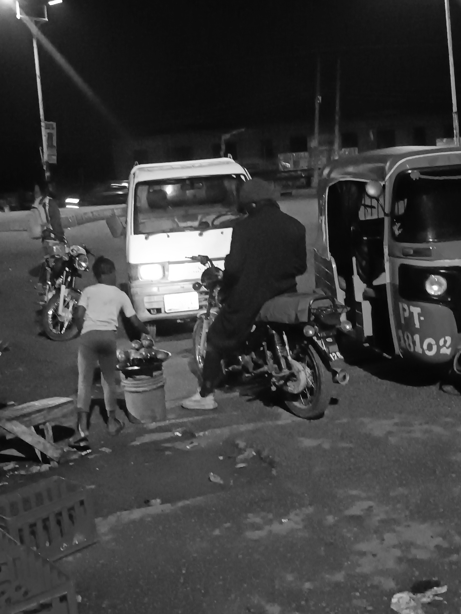 The image captures a scene in Bukuru, Nigeria, at night. A white van is parked between a motorcycle with a seated rider and a three-wheeled motorized vehicle, which displays the registration number "PT-18102." A person wearing light-colored clothing stands near the van, positioned in front of a bucket that appears to hold food items. Another motorcycle is parked nearby. The environment is dimly lit, with a streetlamp visible in the background. Various objects such as pieces of wood and crates are scattered on the ground.