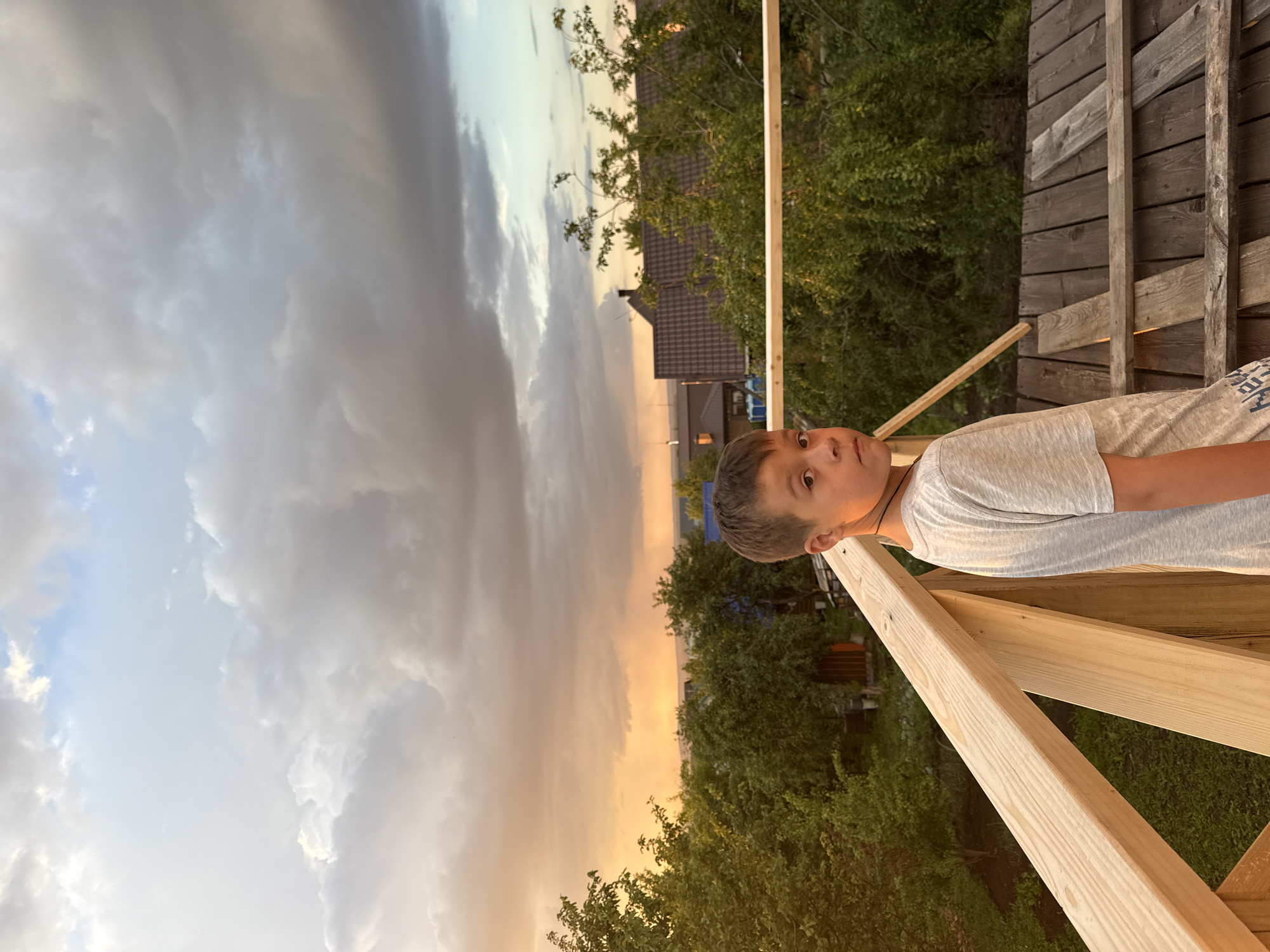 A young boy stands on an unfinished wooden platform, looking over his shoulder at the camera. He is wearing a grey t-shirt. The platform is made of raw lumber and is still under construction. Behind him, lush green trees and the roofs of houses are visible. The sky above is dramatic, with heavy grey clouds tinged with the warm colors of sunset. The scene appears to be in a rural or suburban area, possibly a backyard or a construction site, during the golden hour.