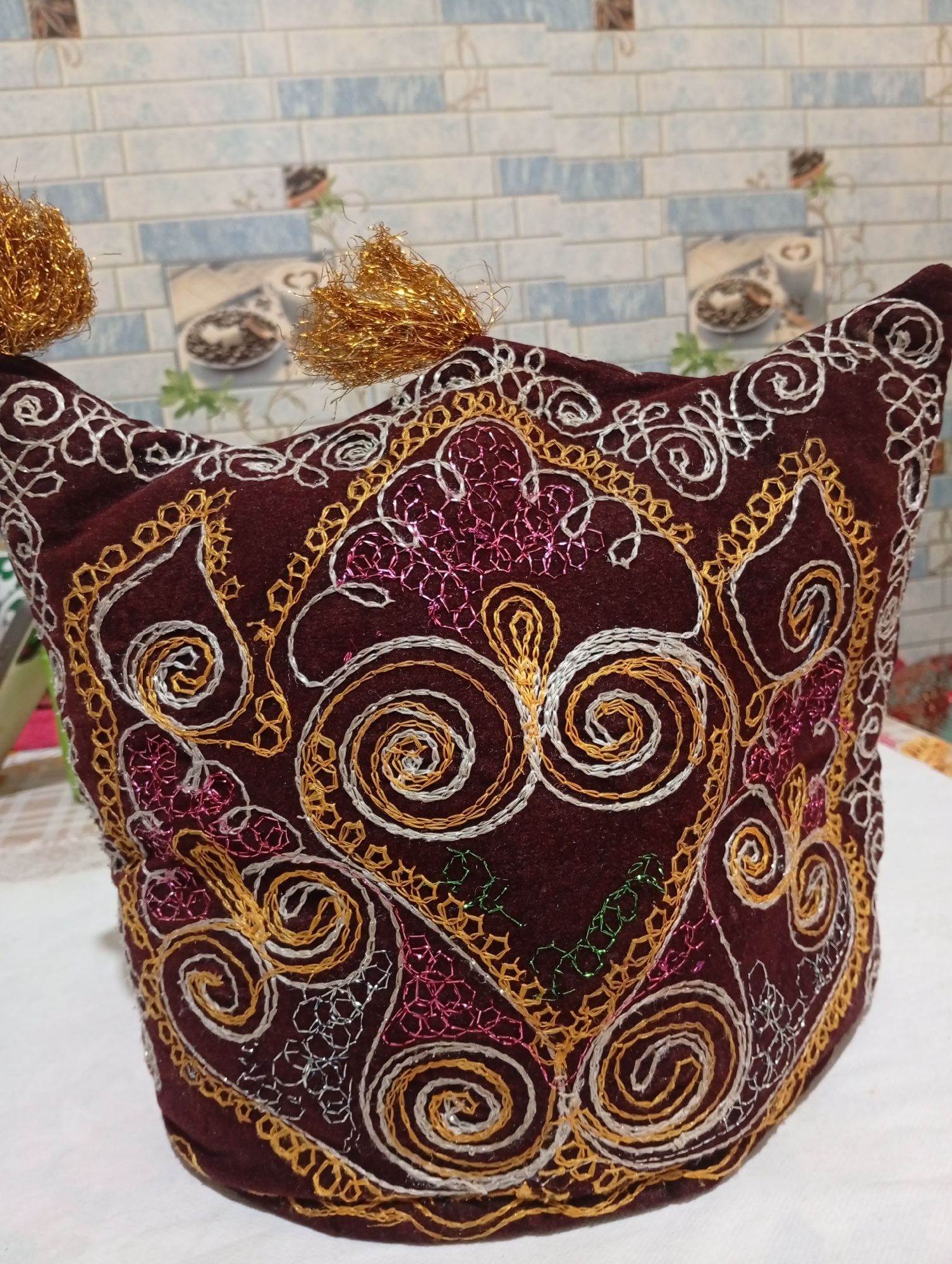 A close-up, eye-level shot shows a richly embroidered, dark red, dome-shaped hat with golden tassels on its upper corners. The hat is adorned with intricate swirling patterns in gold, silver, and pink thread. The background features a tiled wall with a blue and white brick pattern, interspersed with decorative squares depicting coffee cups and foliage. The hat rests on a white surface, likely a tablecloth, with glimpses of other objects on a surface behind it, suggesting an indoor setting. The specific location in Seýdi, Turkmenistan, is not discernible from the image alone.