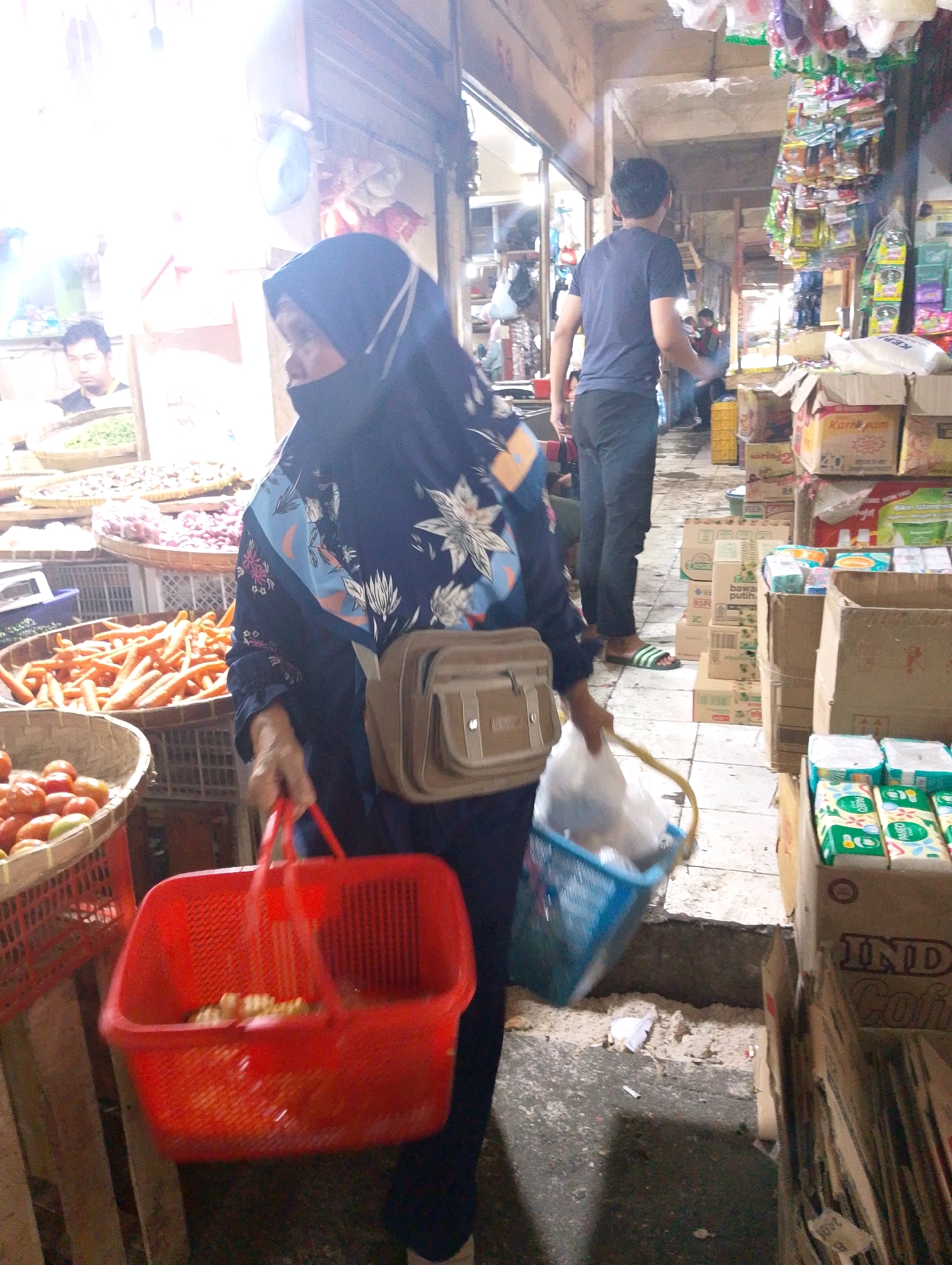 The image depicts a busy marketplace scene in Lembang, Indonesia, likely an outdoor or semi-covered market. In the foreground, a woman wearing a dark hijab and a patterned blue shirt is carrying a large red plastic basket filled with produce. To her right, a blue plastic basket contains a plastic bag. Behind her, a man in a dark blue t-shirt and dark pants walks away from the camera. In the background, stalls are visible, laden with various goods including vegetables like carrots and tomatoes, as well as packaged products such as instant coffee and tissues. The lighting suggests it could be daytime, with some areas brightly illuminated and others in shadow. The ground appears to be paved, with some debris present. The overall impression is one of an active commercial environment with shoppers and vendors.