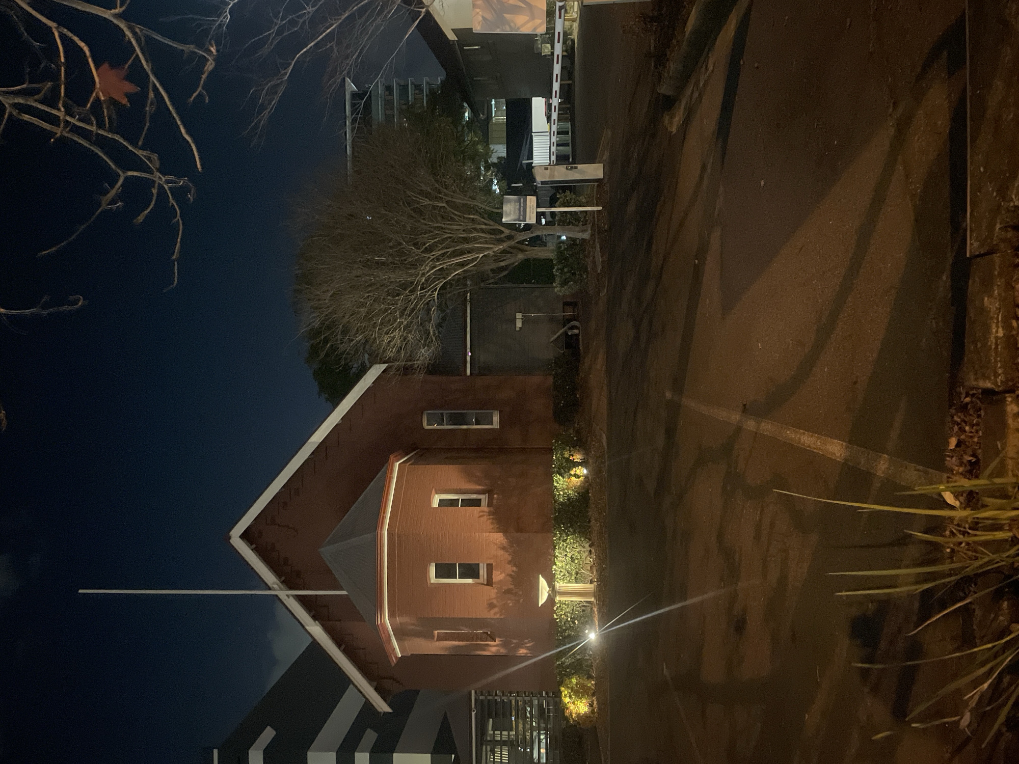 The image depicts the exterior of a small, red brick building with a distinctive octagonal projection at the front, illuminated by ambient lighting at night. A tall flagpole stands against the dark sky, positioned near the peak of the building's roof. The building appears to be part of a larger complex, with a modern, multi-story structure visible to the left, characterized by its dark facade and white horizontal accents. To the right, a gate with a barrier arm is visible, suggesting a controlled access point to the property. Bare trees with spindly branches are scattered throughout the scene, indicating a cooler season. The ground in front of the building is a paved area, possibly a parking lot, with visible white road markings and shadows cast by the lights and structures. In the foreground, some blades of grass and fallen leaves are visible along the edge of the paved area. The overall atmosphere is one of quietness and stillness, typical of a nighttime urban environment. The building's architectural style and the surrounding modern construction suggest it could be a historical building preserved within a contemporary urban setting in Fortitude Valley, Australia.