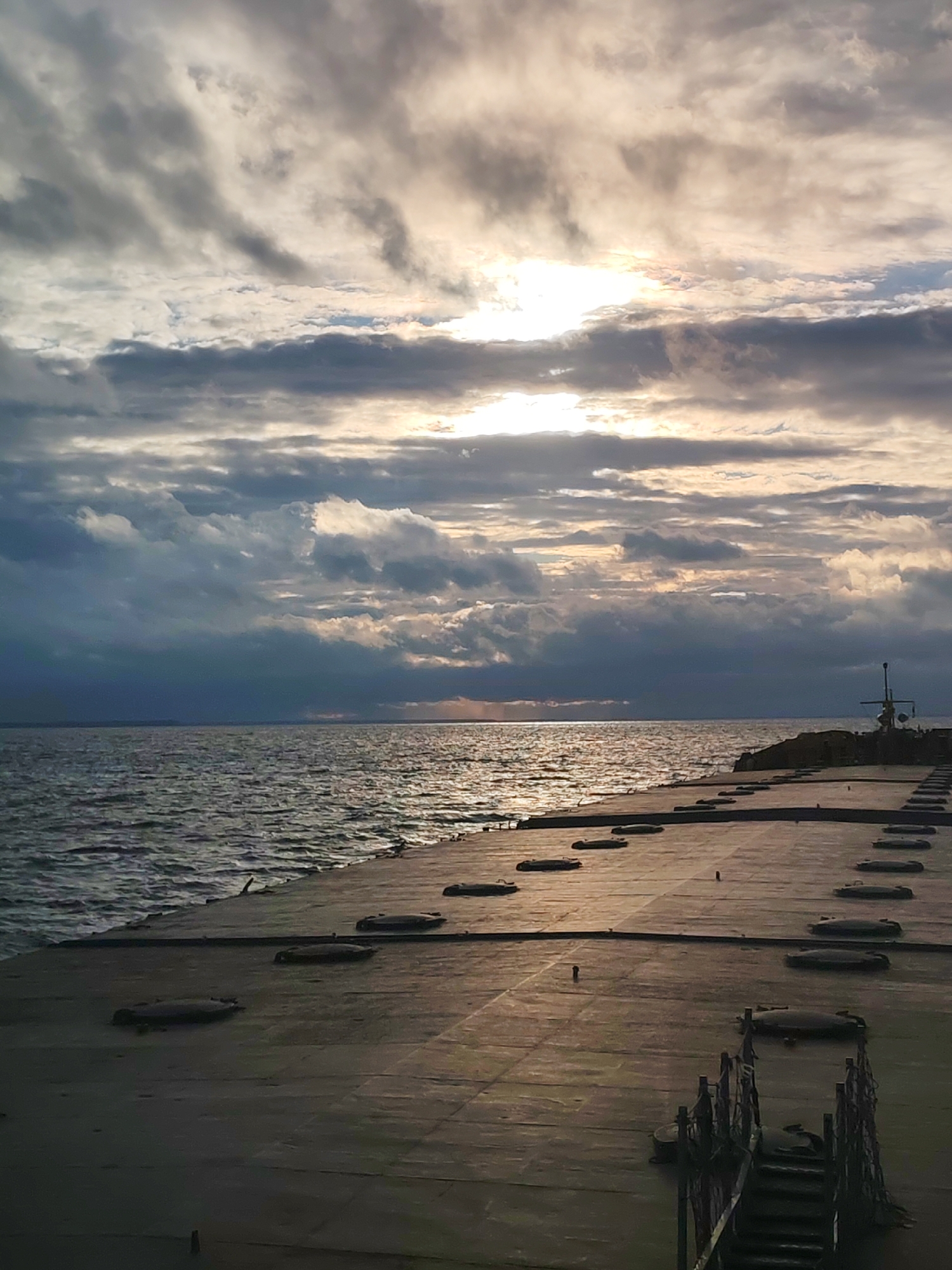 A pier extends into the sea under a cloudy sky. The sun is obscured but visible through the clouds, creating an overall dim, reflective light on the water and pier surface. The pier is concrete with regular, circular indentations. At the end of the pier stands a tall structure. A set of stairs descends from the pier surface, hinting at lower access. The location appears to be a coastal area, but the specific city is not identifiable from the image.