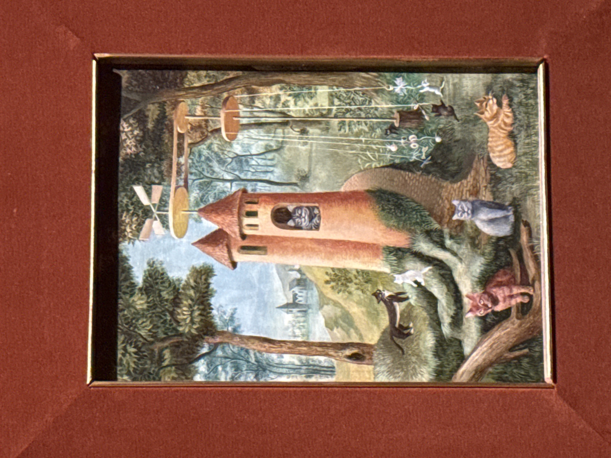 The image is a framed painting. The scene depicts a whimsical landscape with a tall, cylindrical tower structure featuring a window with cats inside. A windmill-like contraption is connected to the tower. Several cats are scattered throughout the landscape, engaging in various activities such as climbing trees and jumping. The background includes trees, foliage, and a distant building. The painting is encased in a dark brown frame with a gold inner lining. There is no indication in the image that it is in Mexico City, Mexico.