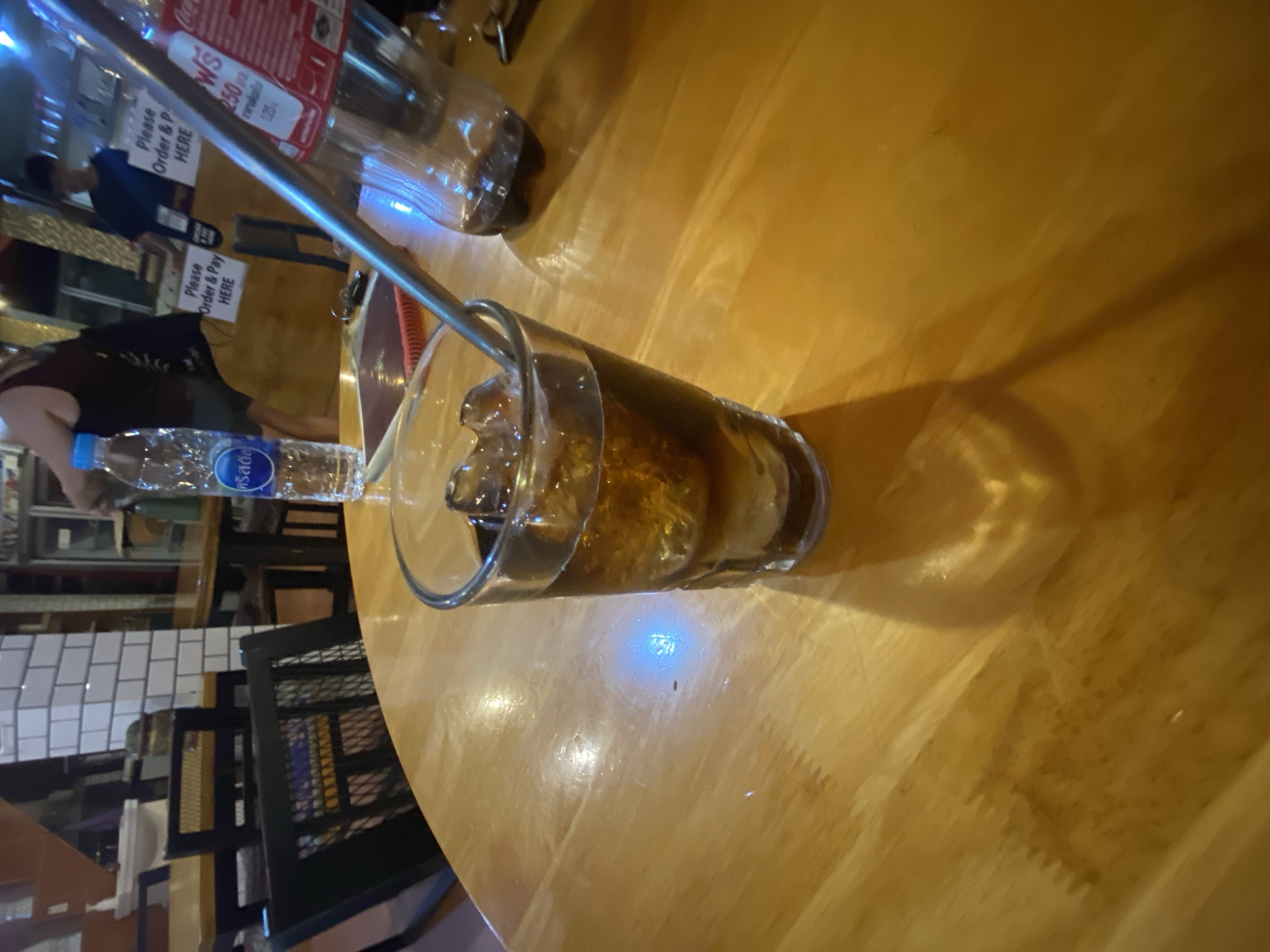 The image depicts a close-up view of a wooden table at a bar or restaurant. On the table, a glass filled with a dark beverage and ice cubes is prominently featured. A metal straw extends from the glass. To the right, a partially visible, empty plastic bottle, likely containing a similar beverage, is present. In the background, a white tiled wall and various restaurant elements, including chairs, tables, and people, are visible. The overall scene suggests a casual dining or drinking establishment. The location is identified as Unknown, Unknown.