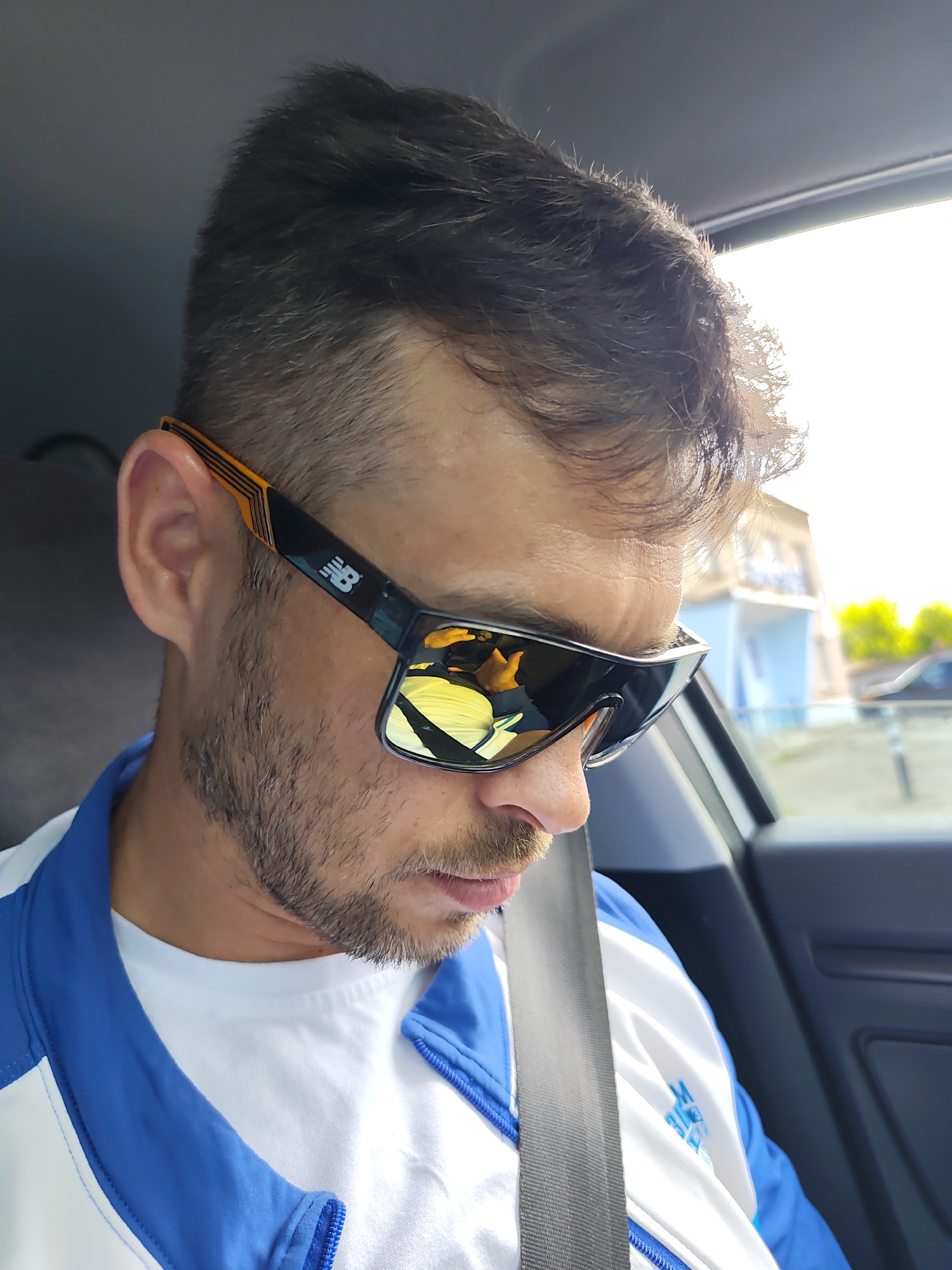 A man wearing New Balance sunglasses, a white t-shirt, and a blue and white jacket is sitting in a car. He is wearing a seatbelt and looking down. Outside the car window, there is a blurred view of a light blue building with a white roof, indicating an urban or suburban setting. The location is likely Chaikovsky, Russia.