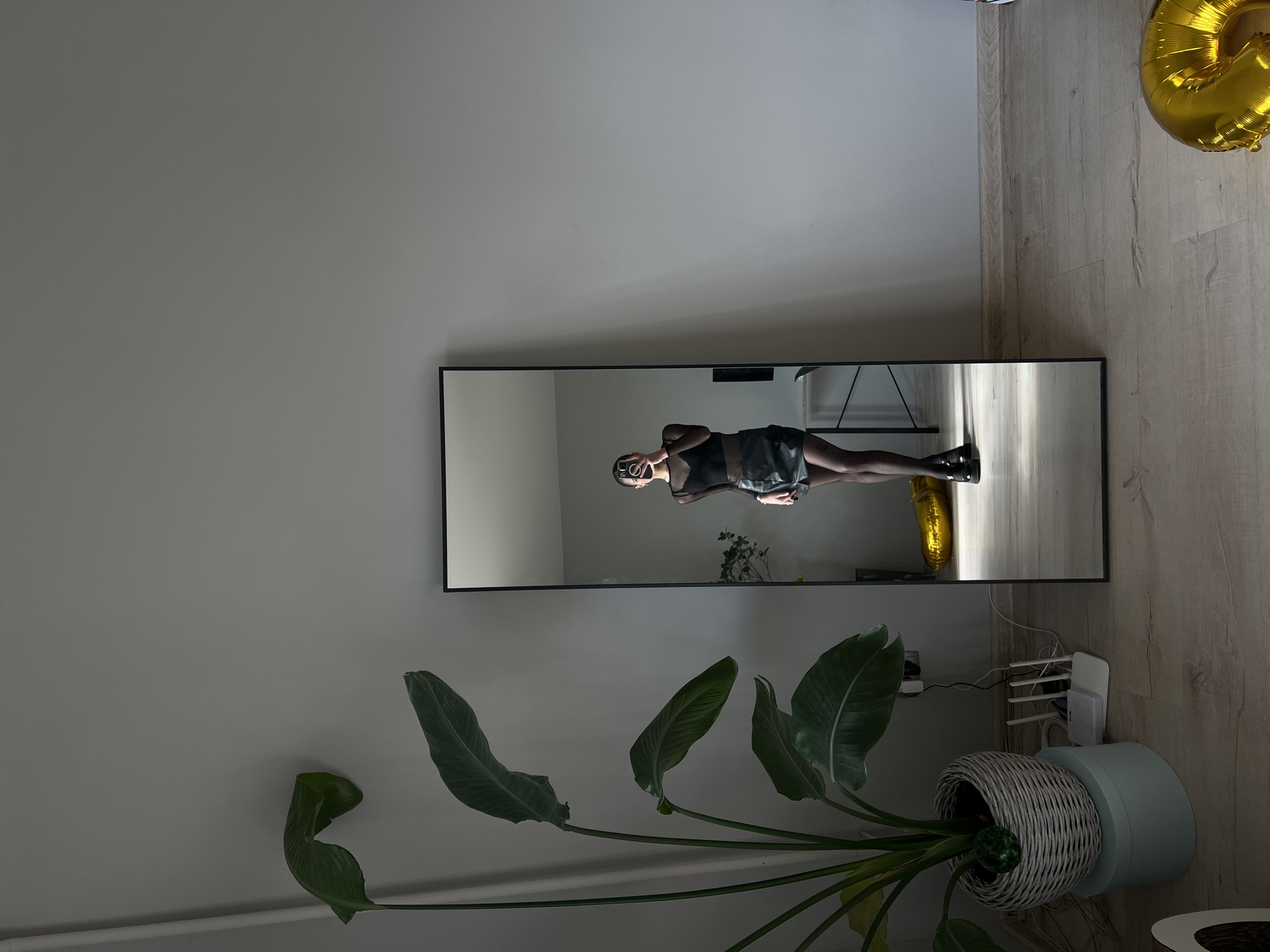 A woman is standing in front of a large, full-length mirror, taking a selfie with her phone. She is wearing a black sheer long-sleeved top, a black leather mini-skirt, black tights, and black shoes. The mirror reflects her entire body and the surrounding room. To the left of the mirror is a large potted plant with wide green leaves. The room has light gray walls and a light-colored wooden floor. In the reflection, a desk with a computer monitor is visible behind the woman. There is also a gold balloon on the floor to the right of the mirror, partially visible. A white router and some wires are on the floor near the plant. The image appears to be taken indoors, possibly in an apartment or studio, and is not explicitly linked to Ternopil, Ukraine.