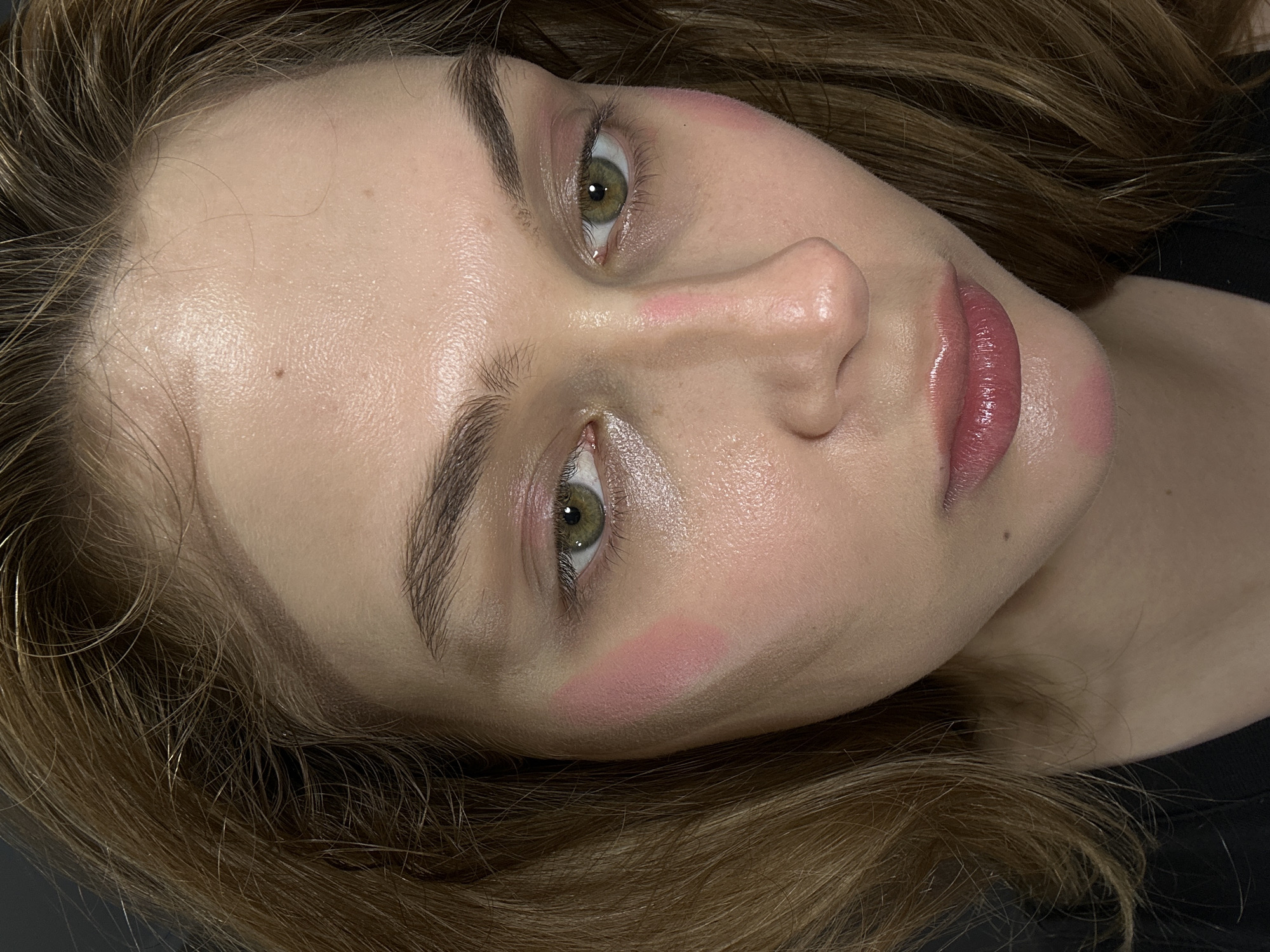 A close-up, slightly tilted, portrait shot of a young Caucasian woman with light brown hair and hazel eyes. She has fair skin and appears to be wearing makeup, including blush on her cheeks and a glossy lipstick. Her eyebrows are well-defined and natural. The background appears to be a dark, possibly black, surface. The image's focus is primarily on her face, highlighting her features and complexion. There is no immediate information to suggest this image was taken in Krasnoyarsk, Russia.