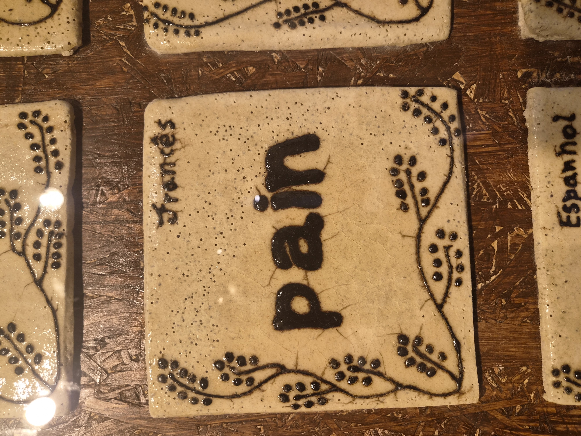 The image displays a close-up, eye-level view of ceramic tiles with dark brown, glossy lettering and decorative elements, placed on a rough, textured wooden surface. The tiles are rectangular and appear to be part of a larger installation, possibly a wall or a decorative piece.
The most prominent tile in the center features the word "pain" written in large, bold, sans-serif letters. To the left of "pain" is a decorative vine-like pattern with small, dark circular berries. Above and to the right of the "pain" tile, another tile shows the name "Francés" written in a smaller, stylized font. Further to the right, partially visible, is another tile with a similar decorative vine pattern. In the bottom section of the image, another tile is visible with the word "Espanhol" and decorative elements.
The ceramic tiles have a matte, speckled off-white finish. The lettering and designs are raised and have a high-gloss finish that reflects light. The wooden background is composed of planks with a visibly rough and splintered texture, suggesting it might be unfinished or distressed wood. The lighting in the scene creates highlights and shadows, adding depth and texture to the image. The overall impression is of a handcrafted, artistic display.
The location of this scene in Seia, Portugal is not indicated by any visual cues in the image itself.