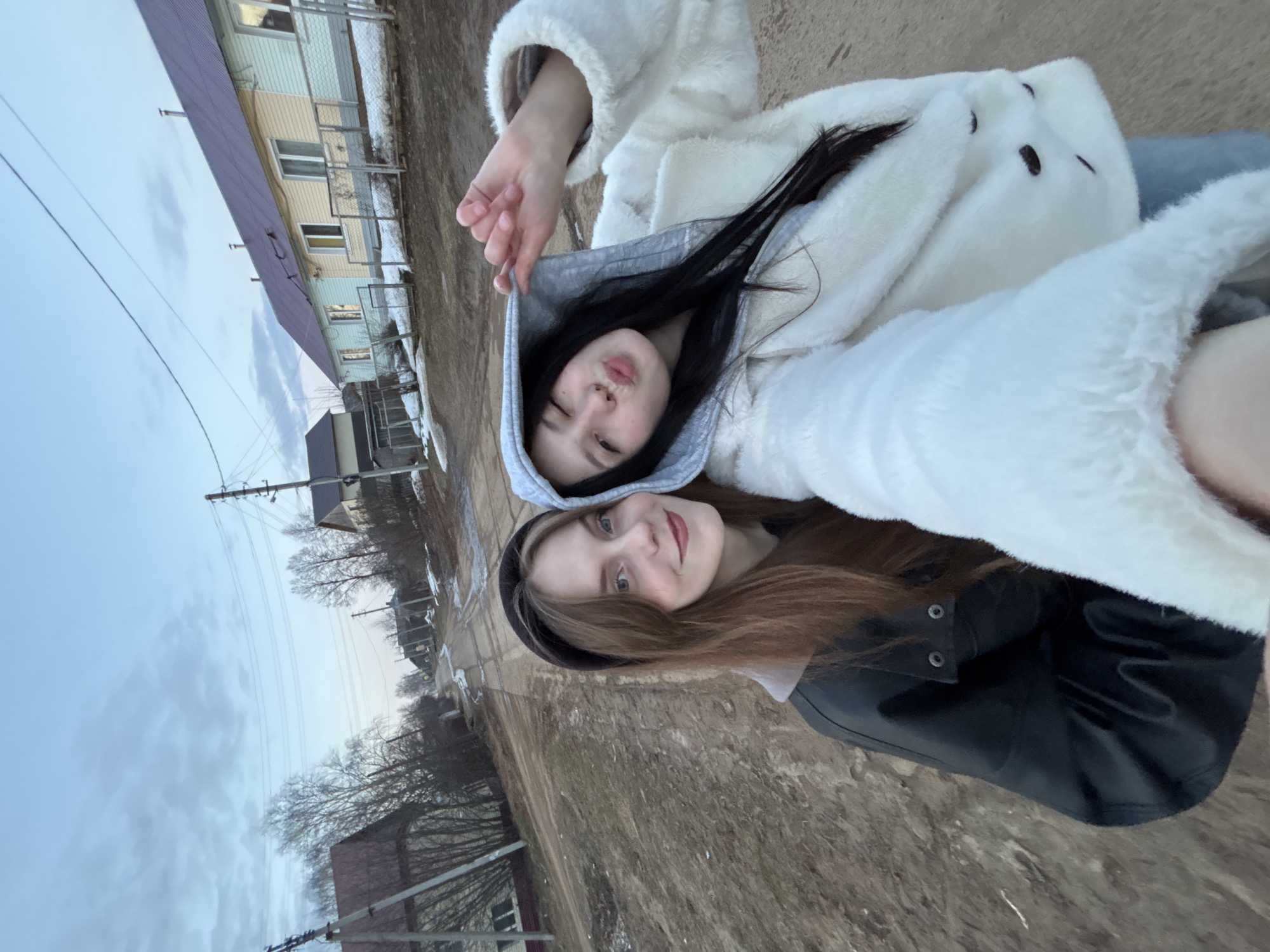 Two young women are photographed outdoors. The woman on the left has dark hair and is wearing a grey hooded sweatshirt under a white, fluffy coat. She has a hand raised near her face and is winking at the camera. The woman on the right has light brown hair and is wearing a black headband and a black leather jacket. They appear to be lying on the ground, possibly taking a selfie. The background includes a building, bare trees, and an overcast sky. The location is Фоки, Russia.