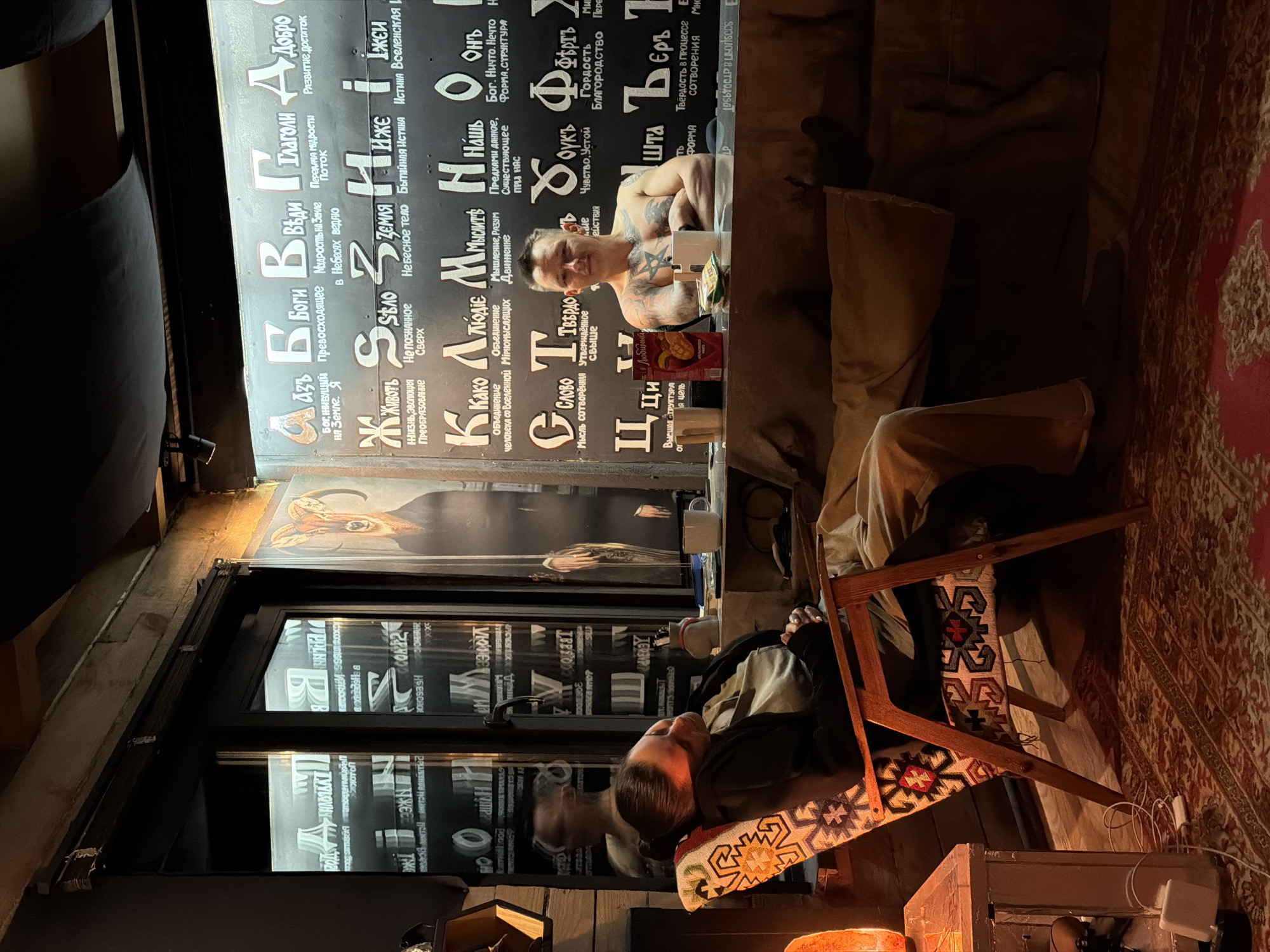The image depicts the interior of what appears to be a themed bar or restaurant, possibly located in Krasnoyarsk, Russia, judging by the Cyrillic text and cultural elements. Two people are visible. One man, heavily tattooed, sits at a table with drinks and snacks. The second person is partially visible, resting in a chair near a window adorned with more Cyrillic inscriptions and a depiction of a ram's head. The environment is dimly lit with a decorative, patterned rug on the floor and an eclectic mix of furniture and decorations, lending an informal and intimate atmosphere to the space. The text displays phrases about being born in the earth or heaven.