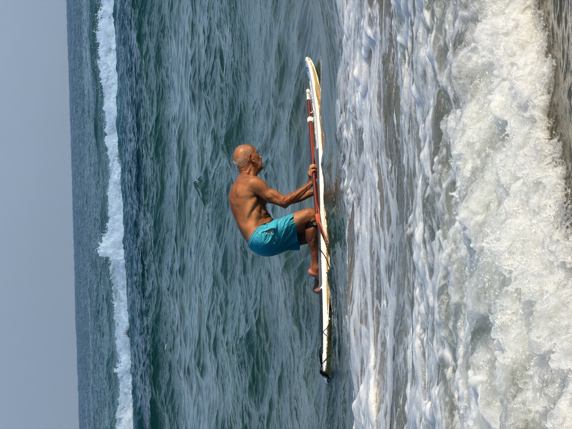 An elderly, bald man with tan skin is kneeling on a surfboard in the ocean. He is wearing blue swim trunks. He is holding a red and white paddle. The surfboard is white with an orange tail. The water is dark blue and choppy with white foam in the foreground. The sky is a light blue with a haze. The image appears to have been taken from a medium distance. The location is the coast of Bat Yam, Israel.