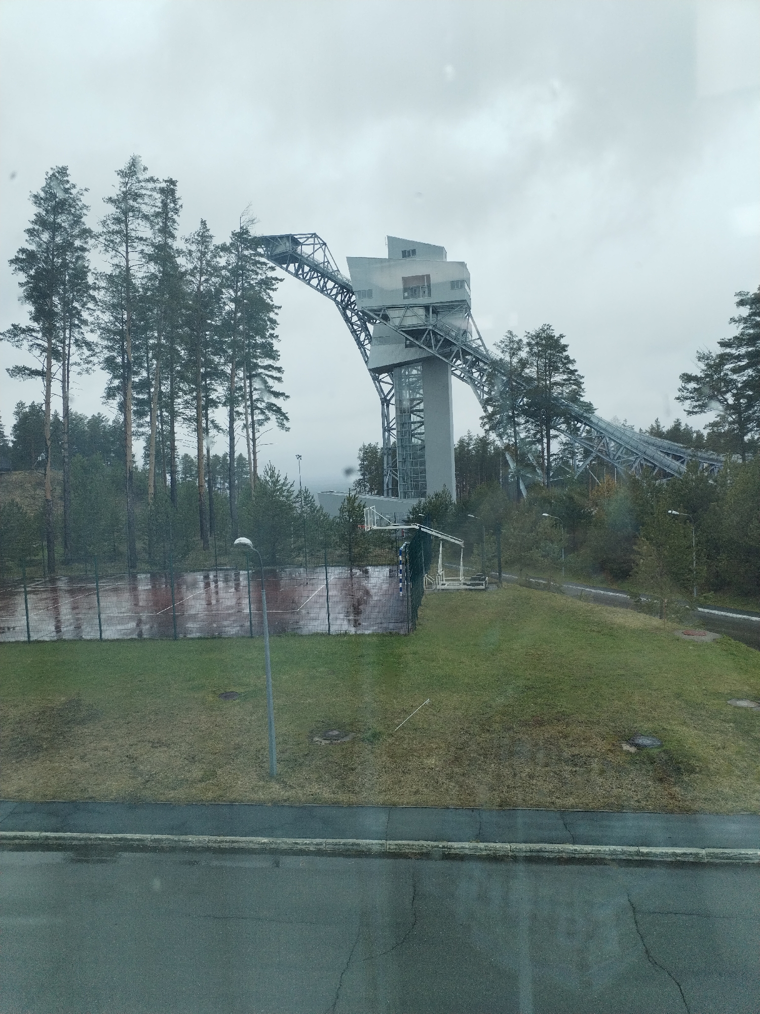 The image depicts a ski jumping hill located in Прикамский, Russia, captured on an overcast day. The steel structure, composed of a tower and descending track, is the prominent element, partially obscured by tall coniferous trees. Below the hill, there is a fenced-in area with a red brick wall visible behind the fence. A grassy slope leads up to the structure, with some patches of brown grass. A lamppost stands on the slope. In the foreground, there is an expanse of paved road with cracks visible. The overall atmosphere is muted due to the grey sky and rainy weather.