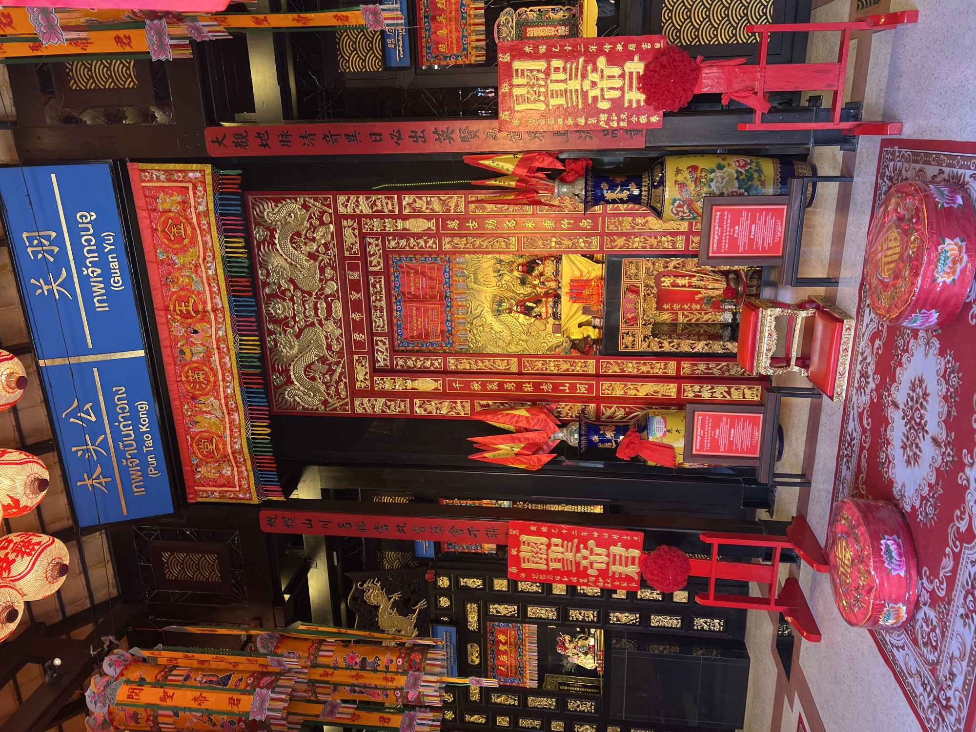 The image shows an indoor shrine in Baan Hua Thanon, Thailand. The shrine features a red and gold altar, adorned with intricate carvings and inscriptions in Chinese characters. Several red objects including small chairs are placed in front of the altar. A blue sign with Thai and Chinese text stands to the left, likely identifying the deities or location. Numerous decorations like lanterns and ornate streamers are visible. The general atmosphere is ornate and culturally significant.