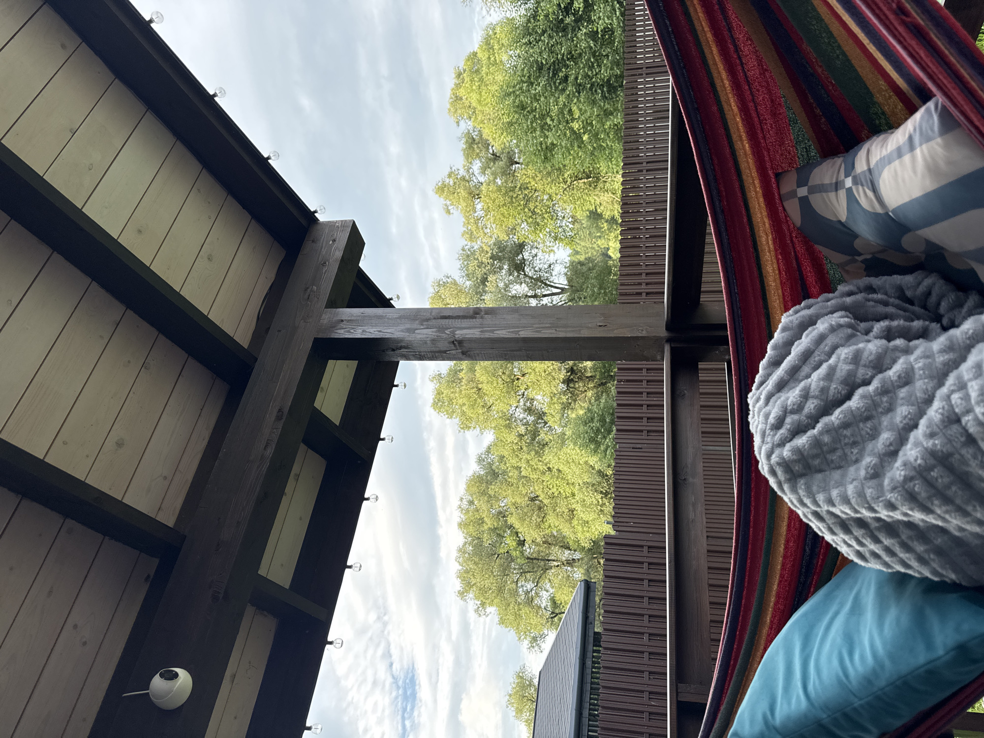 The image depicts a hammock with colorful stripes, adorned with a teal pillow and a gray blanket. A patterned pillow is also visible. The hammock is situated on a wooden deck under a wooden roof, which has string lights attached to the underside. A small white security camera is mounted on one of the wooden beams. In the background, a brown wooden fence encloses a yard with lush green trees and a cloudy sky overhead. To the left, a dark-colored roof of a building is partially visible. The scene appears to be located in the city of Gavshino, Russia, suggesting an outdoor recreational space.