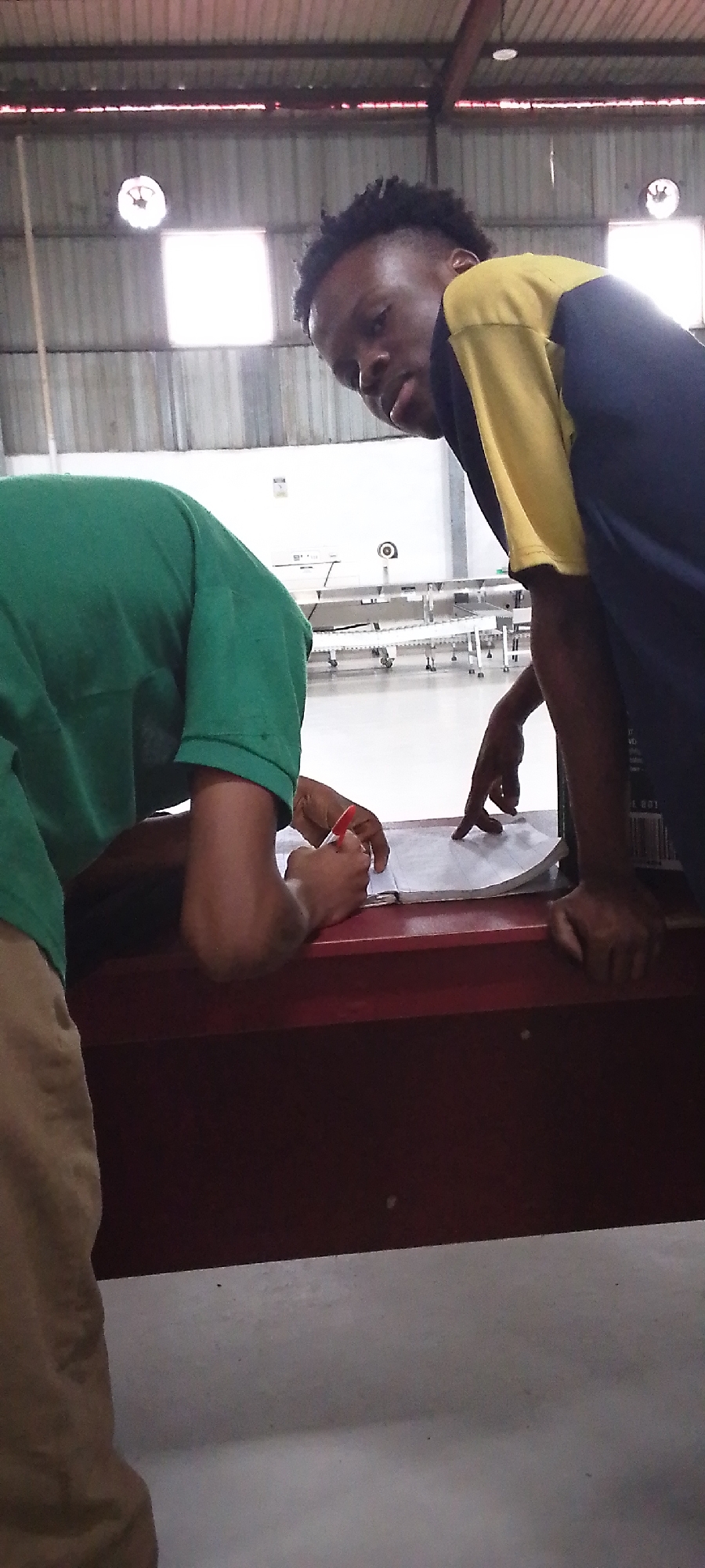 Two individuals are engaged in a work-related activity in what appears to be an industrial or commercial setting. The person on the left is wearing a green shirt and khaki pants, leaning over a red surface and writing on a paper with a red pen. The person on the right is wearing a two-tone blue and yellow shirt, looking directly at the camera while gesturing with their finger on the same paper. The environment includes a metal ceiling with visible light fixtures, a white wall in the background with machinery or equipment partially visible, and a red surface or table where the two are working. The location appears to be in a warehouse or factory of unknown location in the city of Unknown, Unknown.