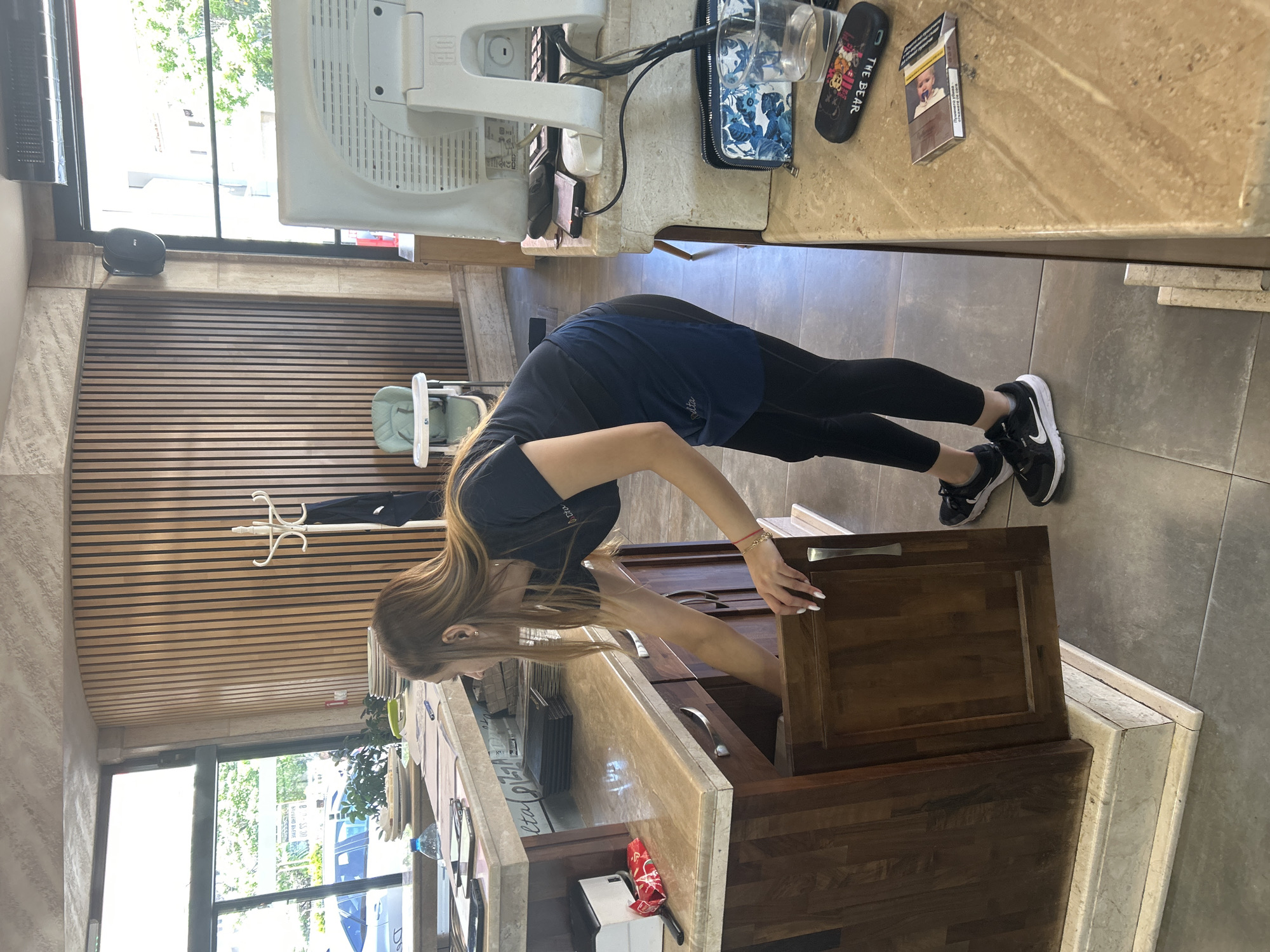 A woman in black athletic wear is opening a dark brown cabinet in a building, likely a shop, in Kazanlak, Bulgaria. The building has a tiled floor and horizontal wood paneling on the walls. A window shows outdoor greenery and buildings. The counter above the cabinet is light brown marble. Items on the counter include a computer monitor, a blue and white floral purse, a phone, and a pack of cigarettes.