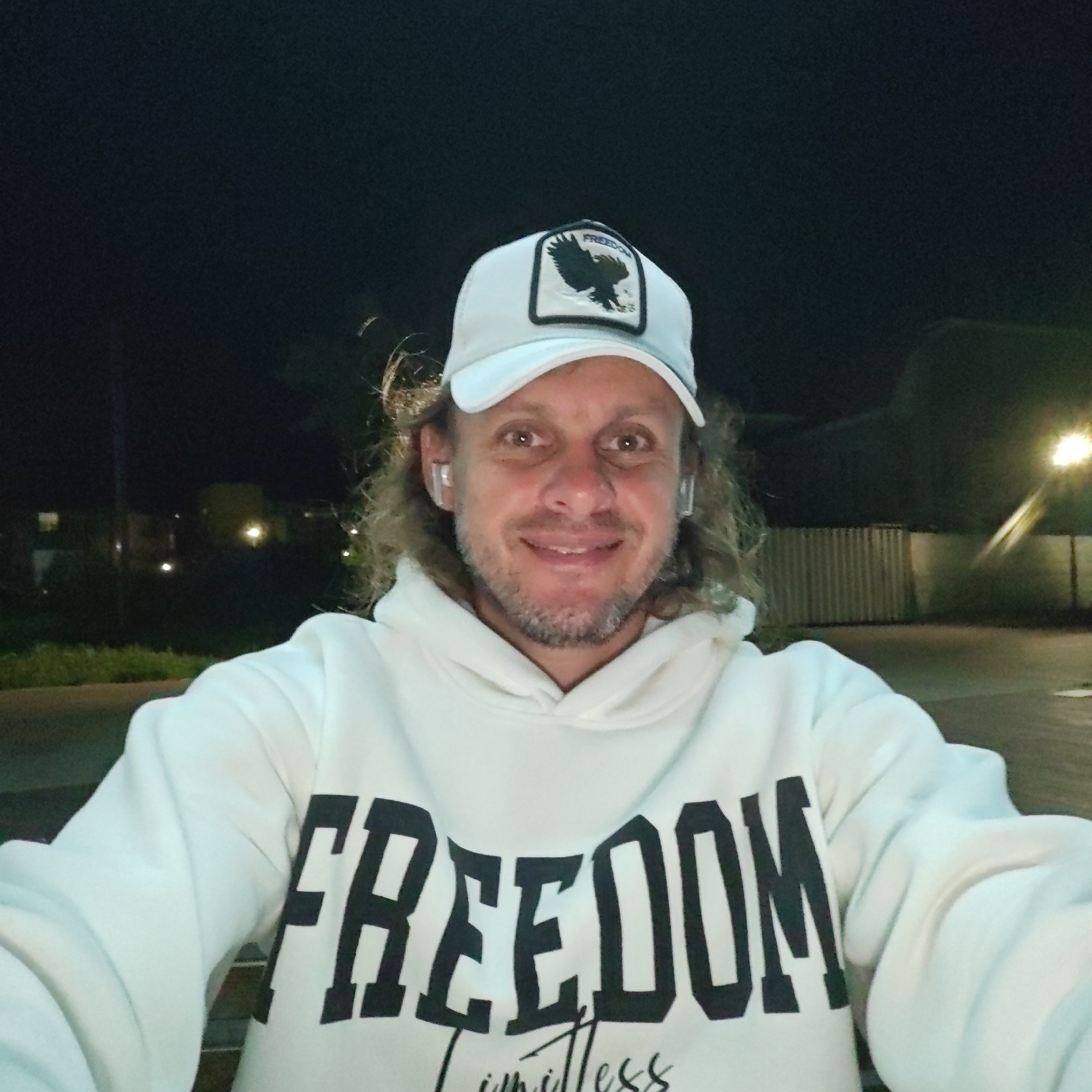 A man is depicted in a close-up, selfie-style portrait taken outdoors at night in городское поселение Щелкино, Ukraine. He has medium-length, wavy light brown hair and a short, light-colored beard, and is smiling directly at the camera. He wears a white baseball cap with a black patch featuring an eagle design; the word "FREEDOM" is visible underneath the eagle on the cap. He is also wearing a white hooded sweatshirt with the word "FREEDOM" printed prominently in large, black block letters across the chest. Below "FREEDOM," the word "Limitless" is written in a smaller, script font. White wireless earbuds are visible in both of his ears. The background is dark, illuminated by blurry artificial lights from what appear to be streetlights and distant buildings. A paved surface and a corrugated metal fence are partially visible in the mid-ground. The time of day is clearly night. The man's expression is happy and relaxed.