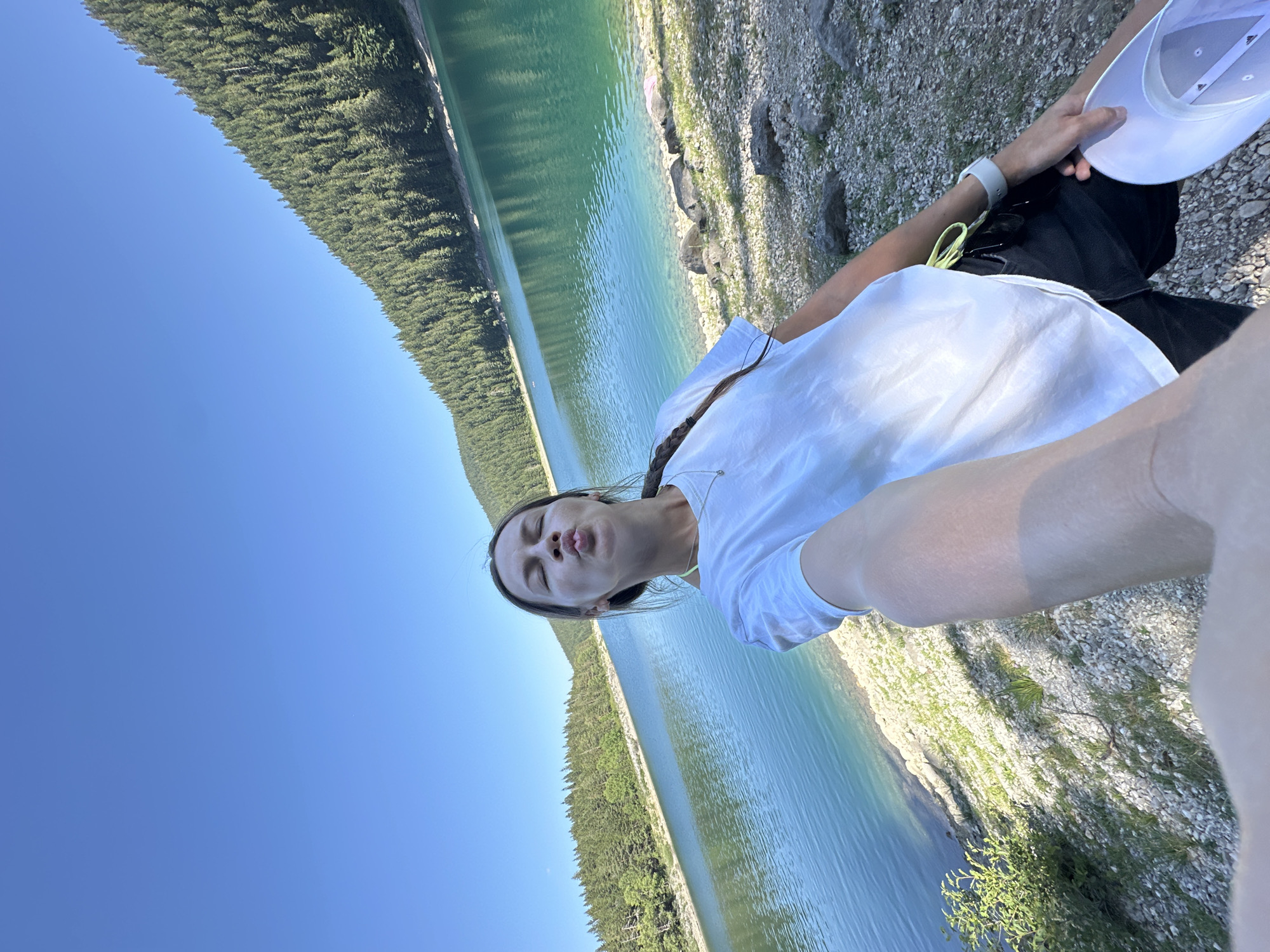 In Pitomine, Montenegro, a young woman with brown hair styled in a braid is taking a selfie outdoors. She is wearing a white t-shirt and black shorts. Her eyes are closed, and her lips are puckered in a kissing gesture, with her tongue sticking out slightly. She is standing on a rocky, grassy embankment next to a clear, turquoise lake. The lake is surrounded by a dense, green forest that climbs up the side of a mountain under a bright blue sky. The woman is holding a white hat in her right hand.