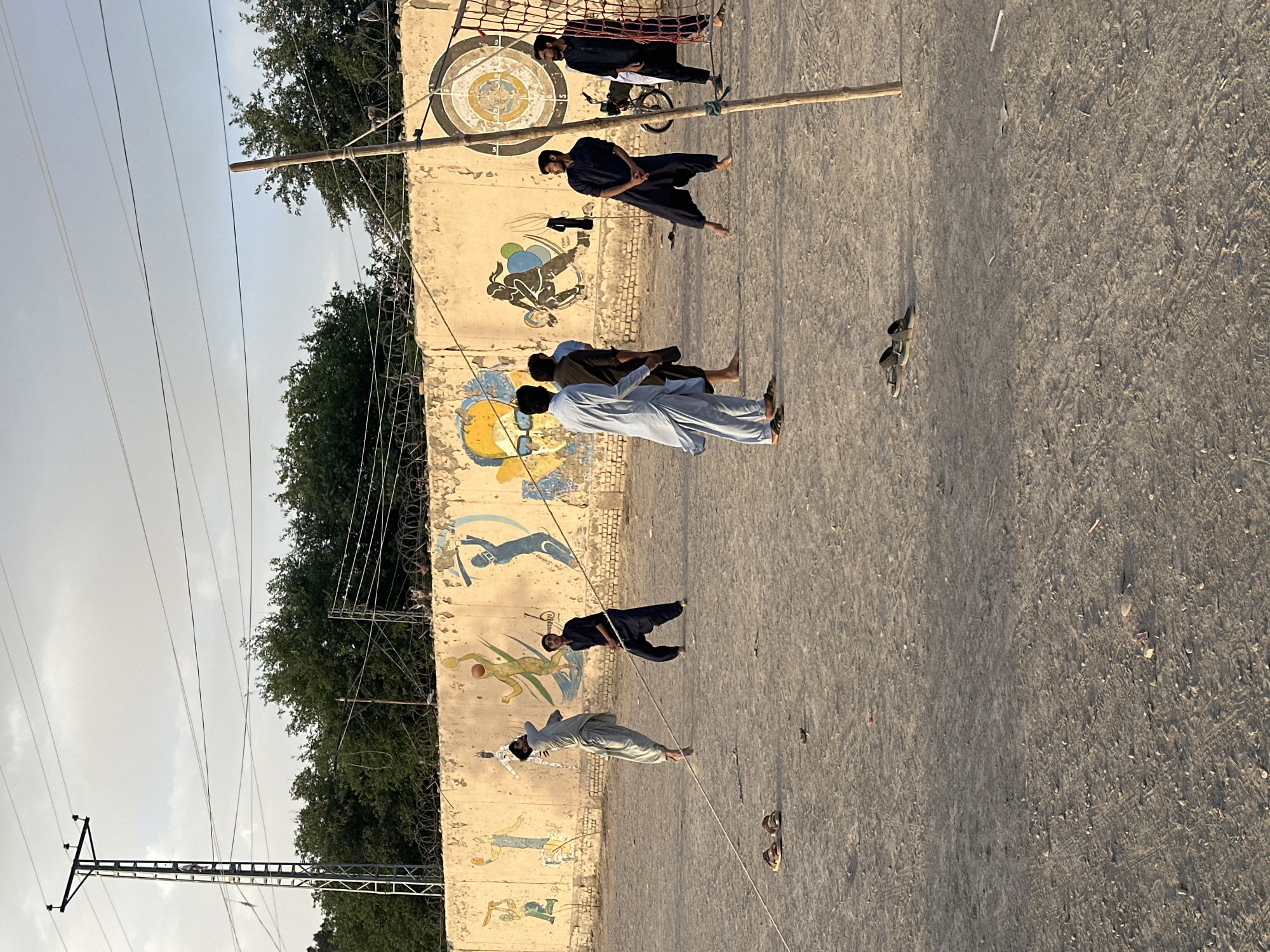 The image shows a group of six individuals standing in front of a long, low wall adorned with faded, cartoonish murals depicting various athletic activities, including a target game. The wall appears weathered and possibly aged, with additional structures like a net and a wooden beam above. The ground is a rough, uneven surface. The sky is a pale, overcast color, and trees are visible beyond the wall. The scene is lit by natural daylight. The photo was taken in Quetta City Tehsil, Pakistan.