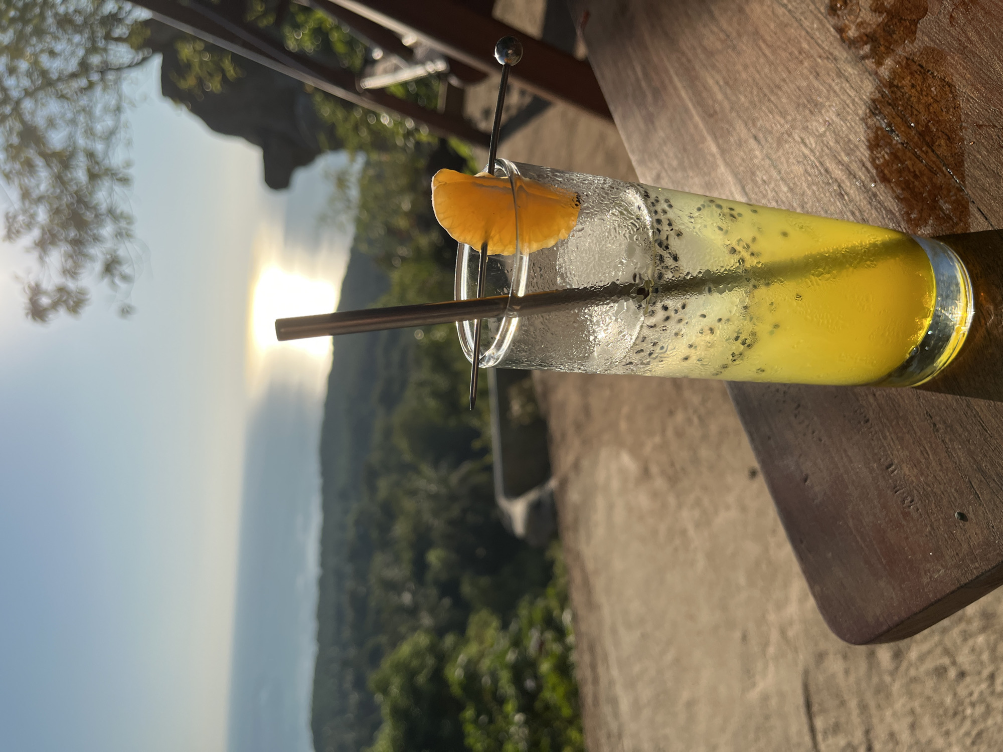 A tall glass of yellow drink with chia seeds and ice sits on a wooden table. A slice of orange adorns the rim of the glass, secured by a decorative metal skewer. A metal straw is inserted into the drink. The glass is condensation-covered. The background shows a hazy view of lush green trees and the ocean, with the sun's rays reflecting off the water. The scene suggests a relaxed outdoor setting, possibly a cafe or restaurant, during the daytime. The lighting indicates it is either early morning or late afternoon, with the sun low in the sky. The overall mood is serene and tranquil, capturing a moment of enjoyment in a tropical location.