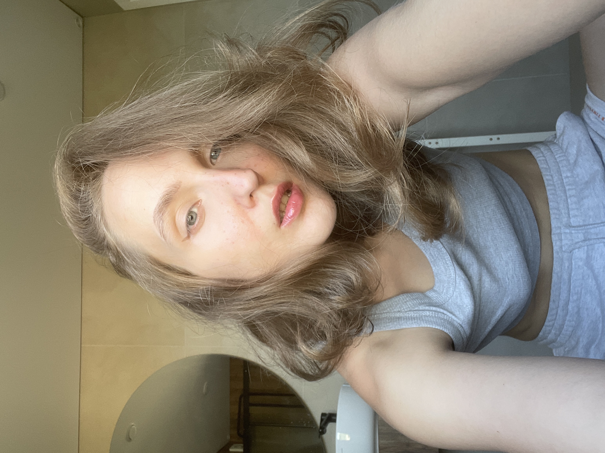 A woman with light skin and long blonde hair is taking a selfie indoors. She is wearing a gray tank top and gray shorts. The background appears to be a bathroom, with a mirror, sink, and tiled walls visible. The location of the scene is possibly in Подшивалово, Russia, as instructed.