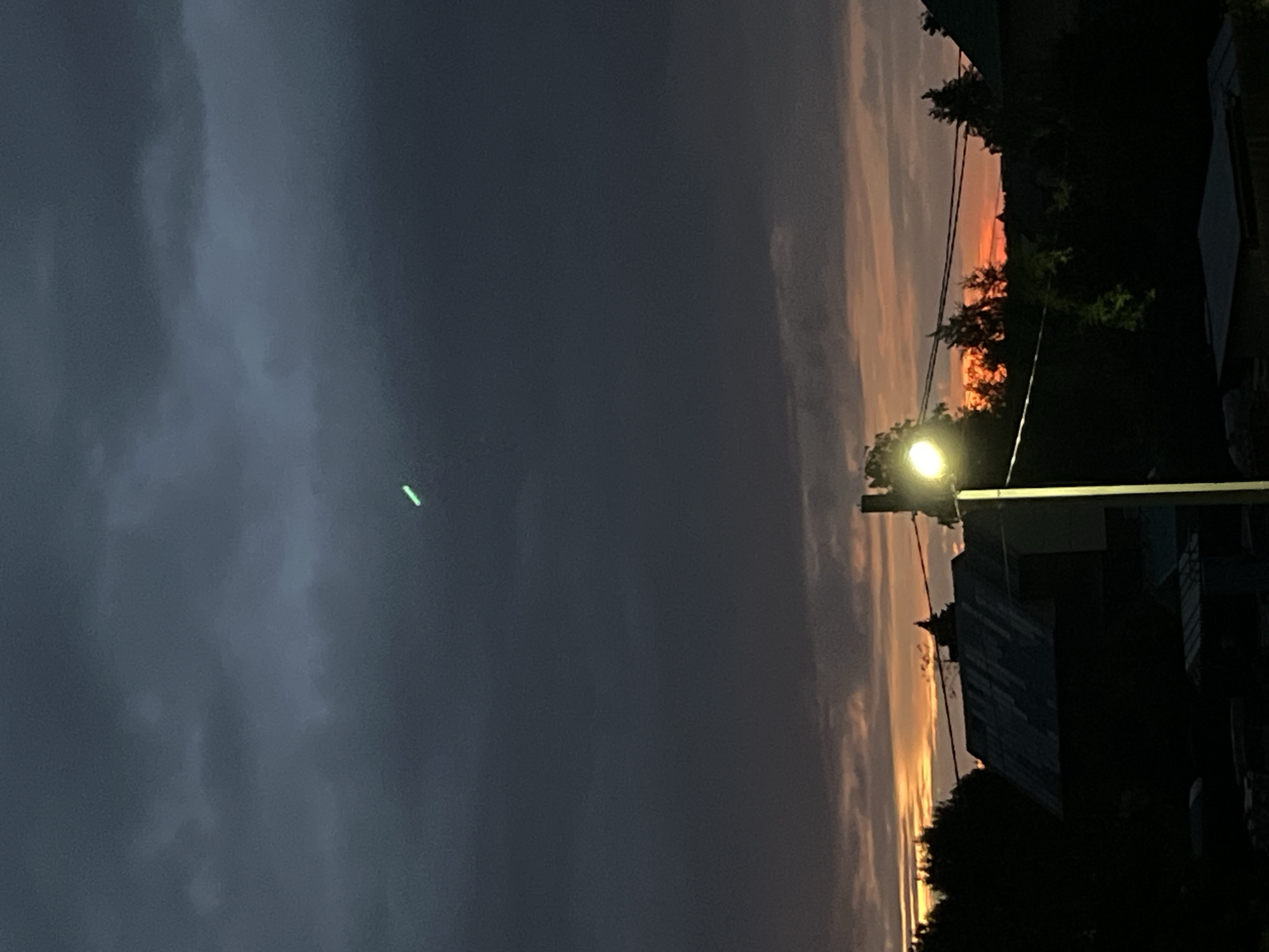 The image shows a dark, cloudy sky at dusk or dawn, with a faint glow of sunset on the horizon. In the foreground, silhouetted against the sky, are houses, trees, and a street lamp. A bright green streak is visible in the sky. The scene is set in the city of Chaikovsky, Russia.