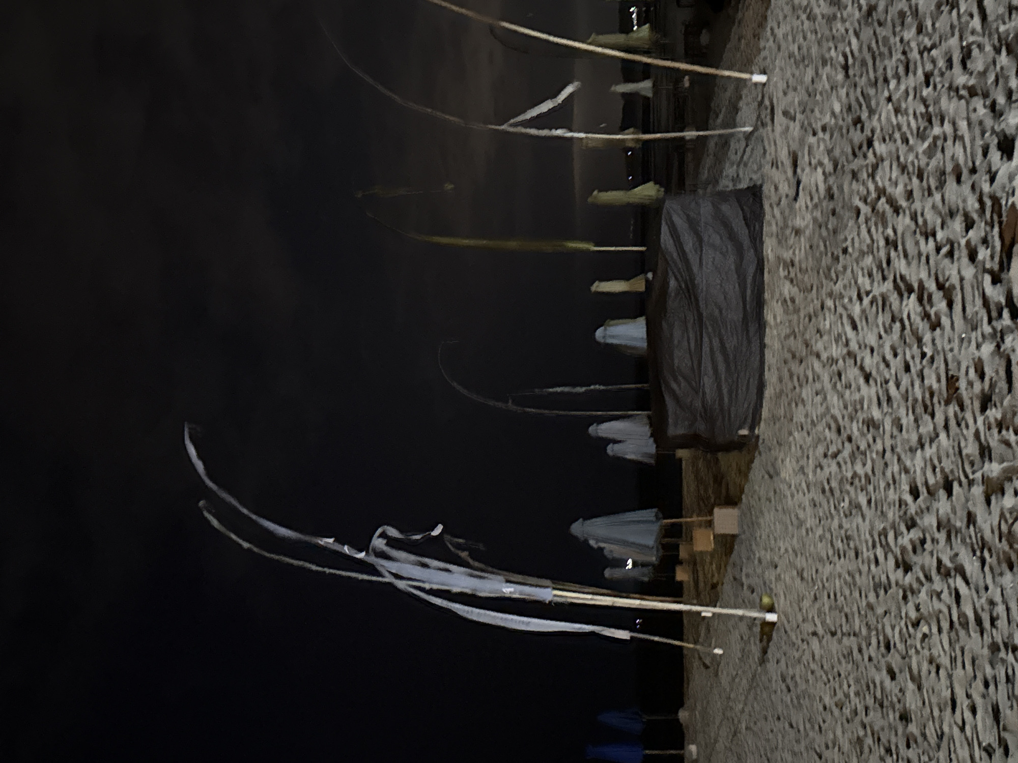 The image depicts a beach at night in Denpasar, Indonesia, likely a public area such as Kuta Beach or Seminyak Beach, known for their vibrant tourist scenes that go quiet after dark. The sand is visible in the foreground, marked with footprints and indentations from the tide. Several beach umbrellas, some closed and covered in light gray fabric, are spaced out across the sand. Tall bamboo poles, characteristic of beach setups, rise from the sand, some with white fabric strips attached, appearing to be decorative flags or wind chimes swaying in the breeze. In the middle ground, a large, dark tarp covers what appears to be a structure or pile of objects, possibly equipment or stored beach furniture. The sky is dark, with faint cloud formations visible, and the overall lighting suggests it is nighttime, possibly illuminated by distant artificial lights or moonlight. There are no people visible in the frame, and the scene appears deserted. The atmosphere is quiet and still, conveying a sense of abandonment or off-season closure.