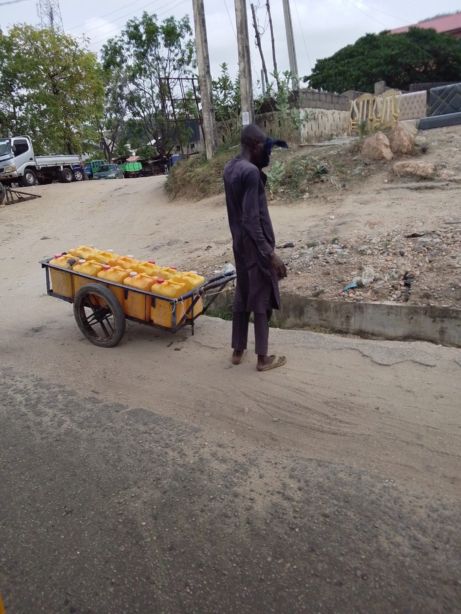 A man is pulling a cart full of yellow plastic containers. The containers are arranged in rows on the cart. The man is wearing a long-sleeved shirt and pants, with sandals on his feet. The cart has a single wheel. The location is a dirt road on the outskirts of Lokoja, Nigeria, with a paved road on the left. Various trees and buildings can be seen in the background.