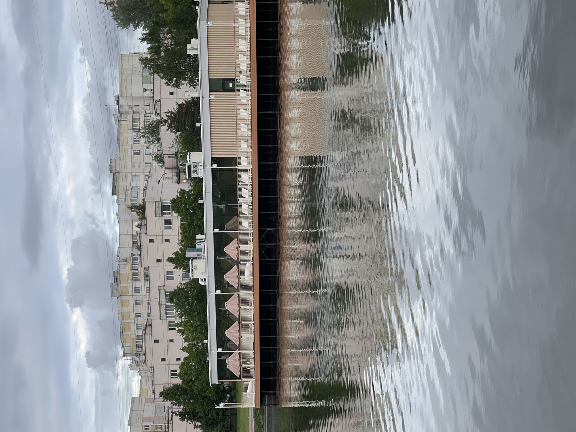 The image shows a waterfront scene in Moscow, Russia, with buildings reflecting in the water. On the left, there are apartment buildings and trees under a cloudy sky. Adjacent to the water is a building with a tan and brown facade, featuring balconies with chairs and umbrellas. The water shows reflections of both the buildings and the sky, with visible ripples. Power lines are visible in the upper left of the image.
