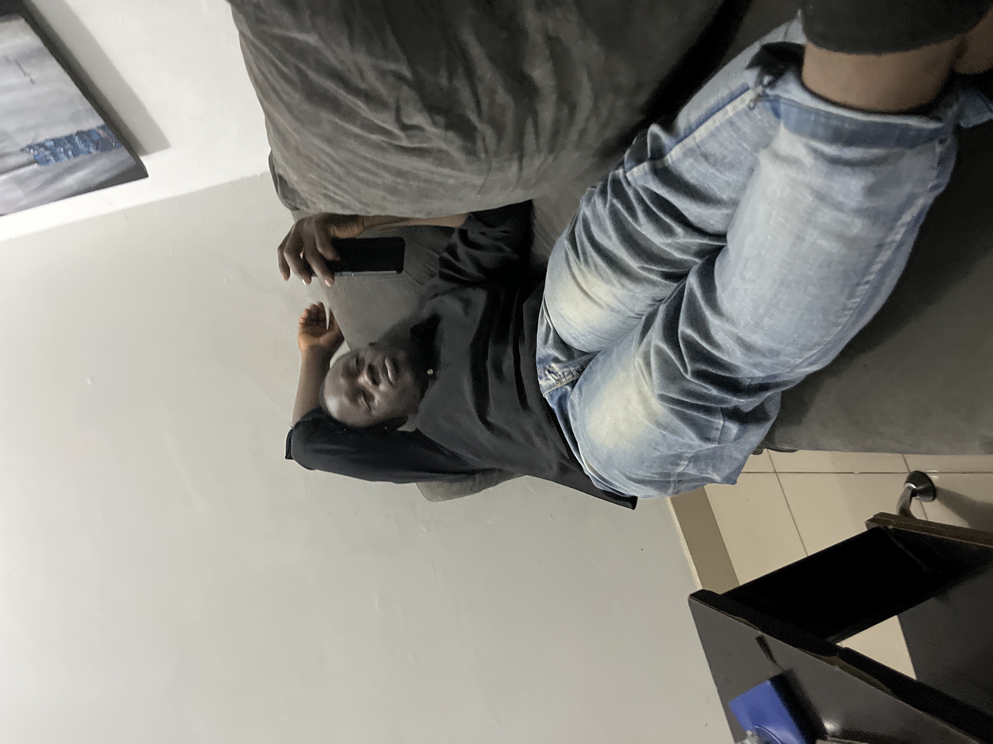 A man is lying on his back on a gray couch with his left arm behind his head. He is wearing a black long-sleeved shirt and blue jeans. He is holding a black smartphone in his right hand. The man's head is tilted back, and his eyes are closed. The couch is in a room with white walls. There is a black table with a blue object on it to the left of the couch. A painting hangs on the wall above the couch. The floor is tiled. The scene appears to be in a residential setting in Abuja, Nigeria.