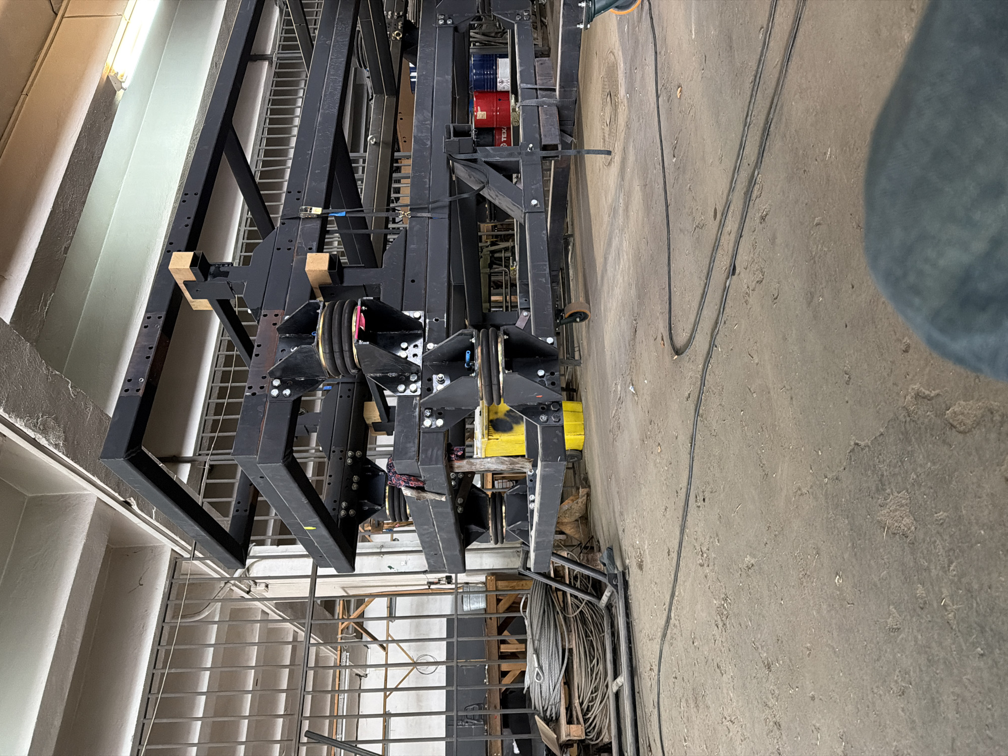 The image shows an indoor industrial setting, likely a workshop or factory. A large black metal structure with multiple pulleys and support beams dominates the center of the frame. This structure appears to be part of a lifting or rigging system. Various tools and equipment are visible in the background, including a ladder, metal shelving, cables, and barrels. The floor is concrete and shows signs of wear and dirt. The image does not offer any details regarding the precise location within Sofia, Bulgaria.