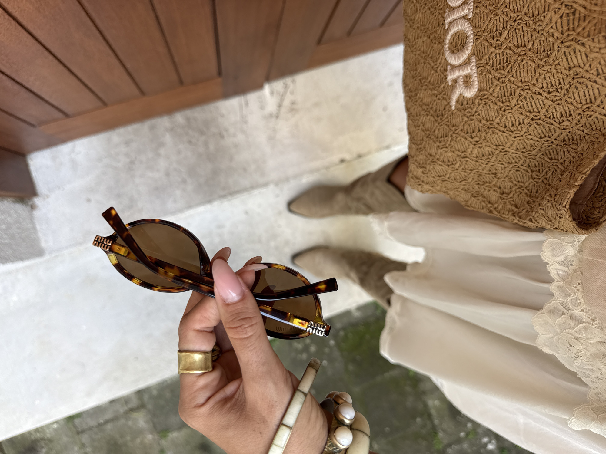A first-person perspective image captures a hand holding sunglasses. The sunglasses are brown tortoiseshell with a distinct logo on the temple. The hand sports a gold ring and multiple bracelets. The individual appears to be wearing a dress or skirt with lace details. A straw bag with the word "DIOR" is also visible. The person is standing on a stone or concrete surface. The image was taken outside with natural lighting. The setting seems to be urban but close to nature, and could possibly be located in North Kuta, Indonesia. The image's focus is on fashion and style.