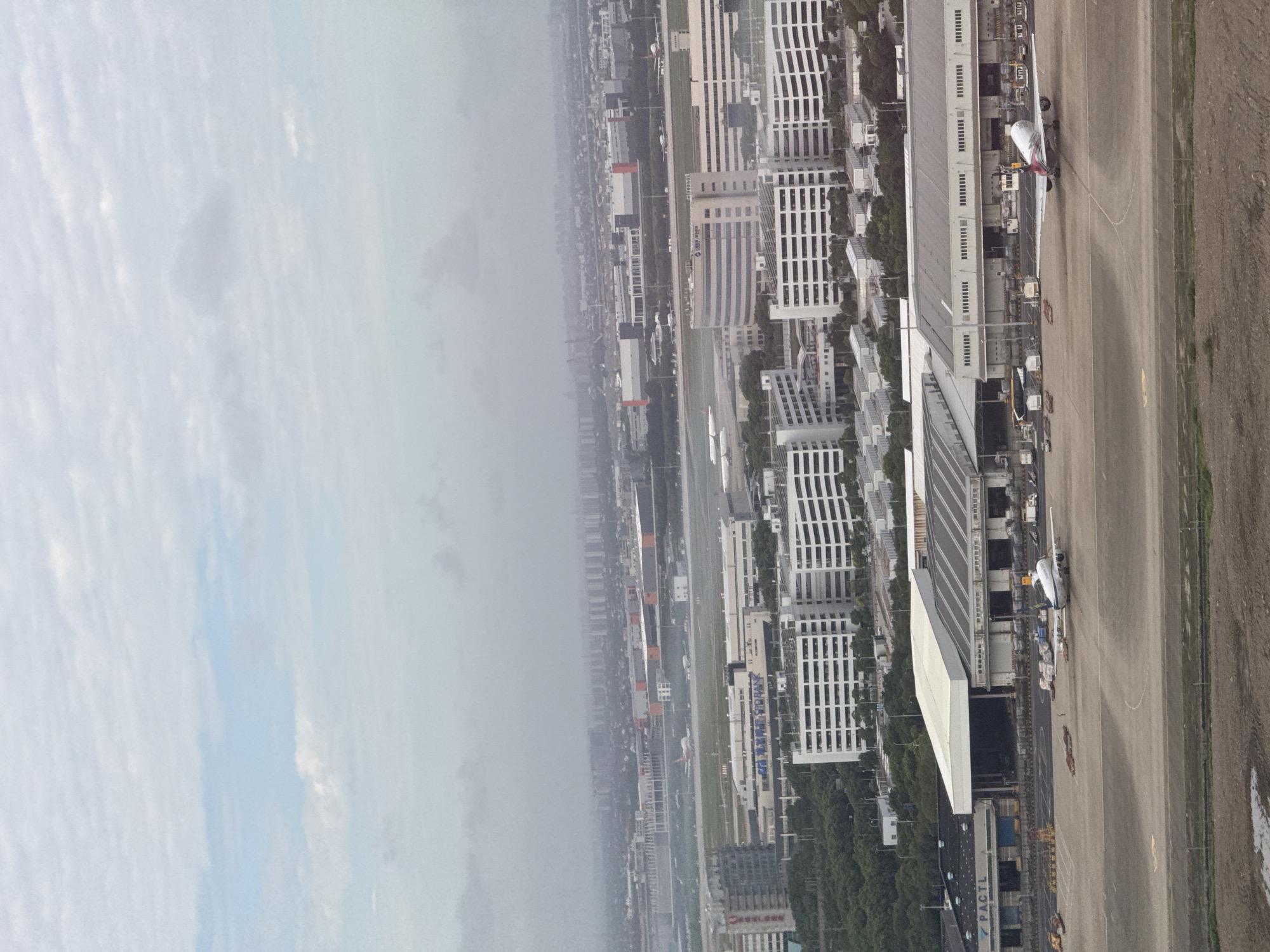 An aerial, long shot shows a cloudy day over Pudong, China, with an airport as the central focus. Large, modern airport terminals and hangars occupy the mid-ground, with multiple airplanes visible on the tarmac. In the foreground, two airplanes are parked on the tarmac. One is a white airplane with red and silver accents, its tail facing the camera. The other, to the left, is mostly white with some red on its tail and is seen from the side. Several vehicles, including what appears to be a tug and baggage carts, are scattered around the parked aircraft. Beyond the airport, a sprawling urban landscape of low-rise and high-rise buildings stretches into the hazy distance, with a dense layer of clouds obscuring the horizon. The overall impression is one of industrial activity and urban expansion under a muted sky.