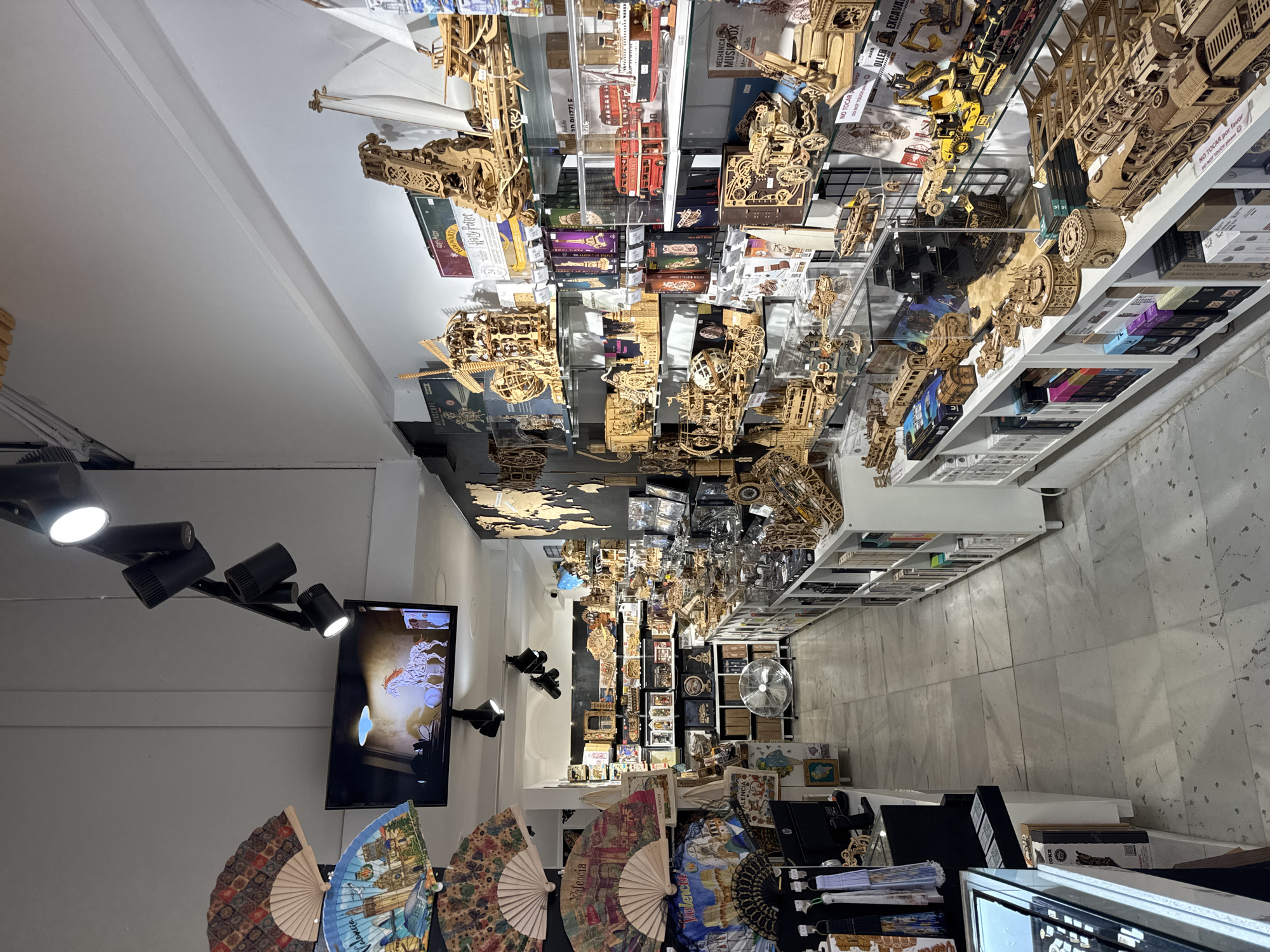 The image depicts the interior of a retail store specializing in wooden model kits, likely located in Valencia, Spain, based on the presence of Spanish text and imagery on the decorative fans. The store is filled with shelves displaying a wide variety of intricate wooden models, including trains, cars, ships, buildings, and mechanical puzzles. The lighting appears to be a mix of track lighting and ambient store lighting, illuminating the merchandise. On the left side of the image, decorative fans with imagery of Spanish landmarks, including what appears to be a depiction of Valencia, are displayed prominently. A television screen is mounted on the wall, showing a moving image of a horse. The floor is tiled with a light-colored marble pattern. Several customers are not visible in the image. The overall atmosphere suggests a shop catering to hobbyists and collectors.