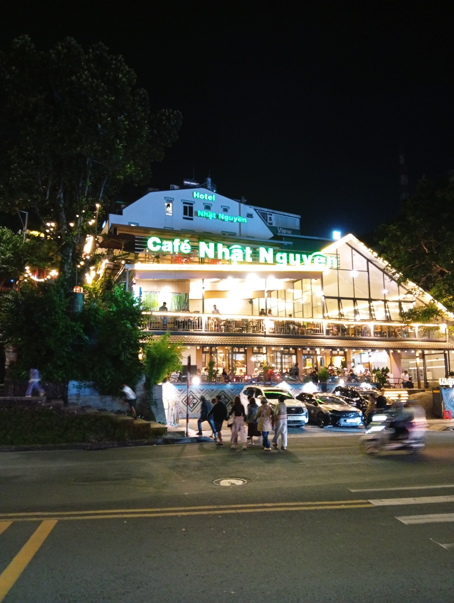 The image depicts a street scene at night in Da Lat, Vietnam, with "Cafe Nhat Nguyen" as a focal point. The cafe, featuring two stories and illuminated by bright lights, has large windows and balconies with patrons seated at tables. Above the cafe, there is a building labeled "Hotel Nhat Nguyen View". Street level shows pedestrians crossing the road in front of parked cars and a passing motorcycle. The road has visible yellow lines marking lanes and crosswalks. A large tree with string lights adds to the ambiance on the left side of the frame. In the distant background, a tall structure is visible on the right side.