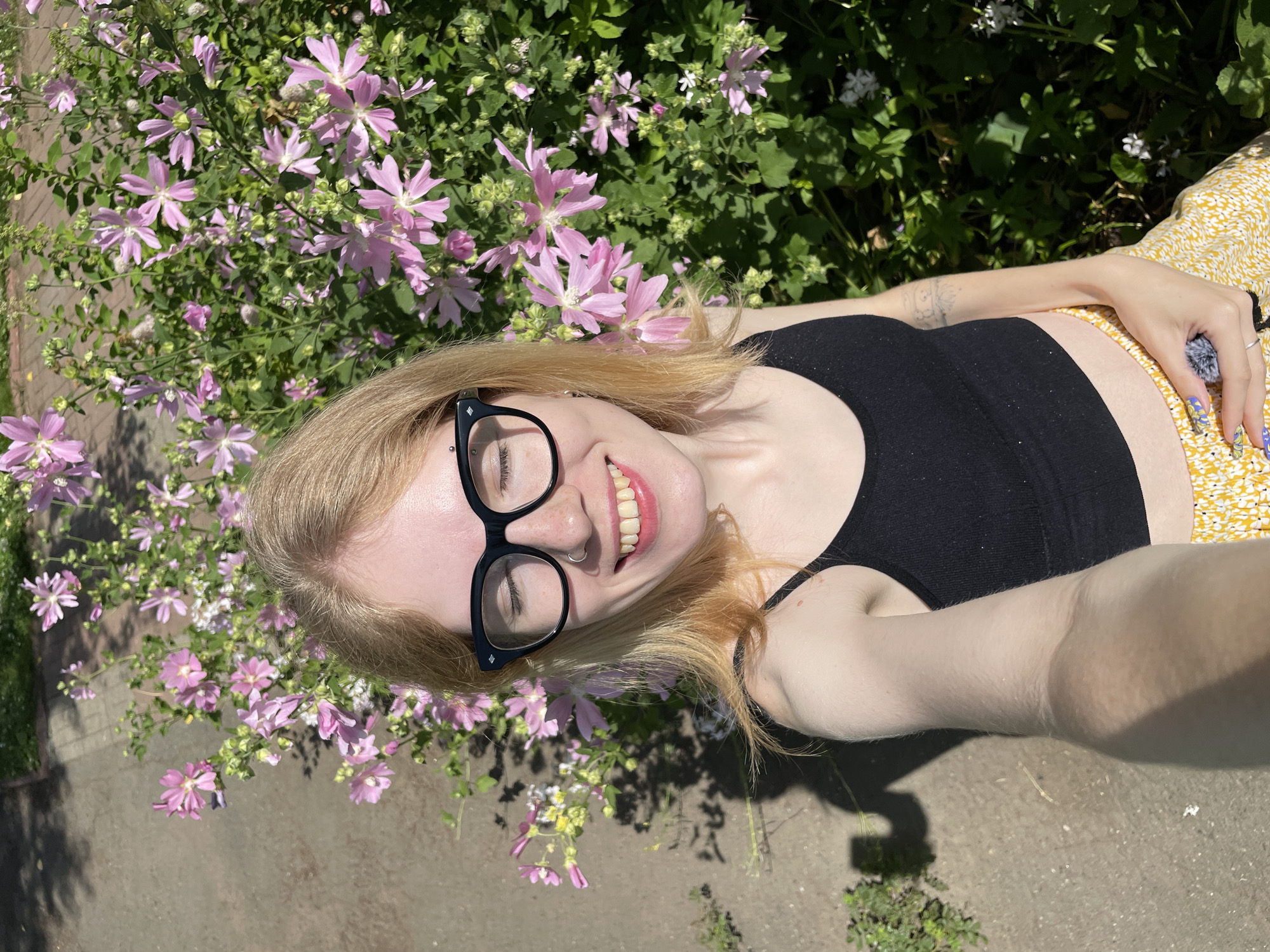 The image is a selfie taken outdoors on a sunny day, likely in Izhevsk, Russia, given the context. The subject is a young woman with blonde hair, wearing black-rimmed glasses and a black tank top. She has a nose ring and is smiling with her eyes closed. She is positioned in front of a lush green bush with numerous pink flowers in bloom. The background also shows a paved walkway and shadows cast by sunlight. The woman is holding a small, fluffy object in her right hand. Her left arm is resting on her lap, revealing a tattoo on her forearm and a yellow skirt with a floral pattern. The overall scene suggests a casual, outdoor setting, possibly a park or garden.