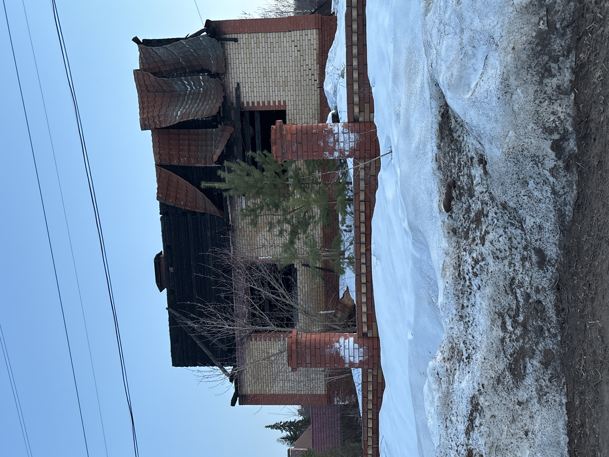 The image shows a fire-damaged brick house, likely in the aftermath of a fire. The house's roof has partially collapsed, with charred remains visible. The brickwork of the house appears to be a mix of light and dark colors, suggesting different stages of construction or damage. There is snow on the ground and a partially brick fence around the house, which indicates the location is likely in a cold climate during winter or late winter/early spring. There are power lines are visible, and the location is described as Chaikovsky, Russia. No people are visible.