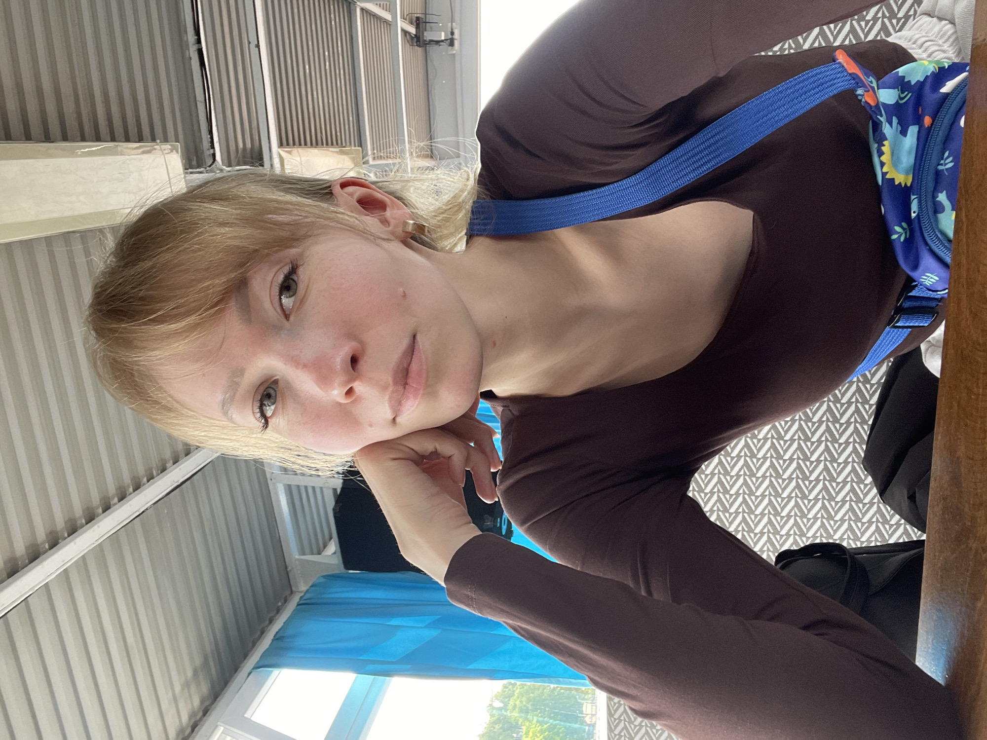 A young woman with blonde hair, fair skin, and light-colored eyes is captured in a close-up, angled portrait, likely a selfie, in Perm, Russia. She wears a form-fitting, long-sleeved dark brown top and has a blue strap crossing her body, which connects to a small blue bag featuring colorful dinosaur patterns in blue, yellow, and green. A gold-colored ear cuff is visible on her left ear. She is seen resting her chin on her right hand, gazing calmly and directly at the camera with a gentle expression.
The setting appears to be an indoor space, possibly a cafe or waiting area, illuminated by bright natural light, indicating it is daytime. The background shows light gray corrugated metal panels on what seems to be a wall or ceiling. A window on the lower left reveals green trees outside. Below the woman, a light gray and white geometric patterned surface is visible, along with a part of a dark bag and a wooden surface to her right. There is no visible text in the image.