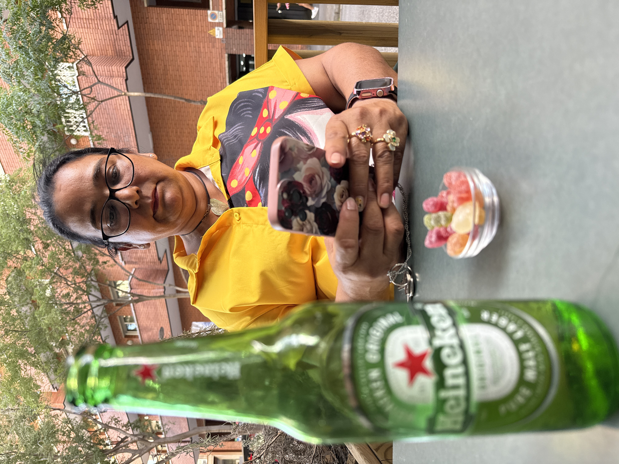 A woman with dark hair and glasses is sitting at a table outdoors, looking at her phone. She is wearing a bright yellow shirt with a colorful, graphic print on the front. In the foreground, a green bottle of Heineken beer is in focus, with its label clearly visible. To the right of the bottle, a small glass bowl contains several colorful gummy candies. Behind the woman, a brick building with windows and trees can be seen. The scene appears to be a casual outdoor setting, possibly a cafe or restaurant terrace in Barcelona, Spain, given the style of the building and the presence of outdoor seating.
