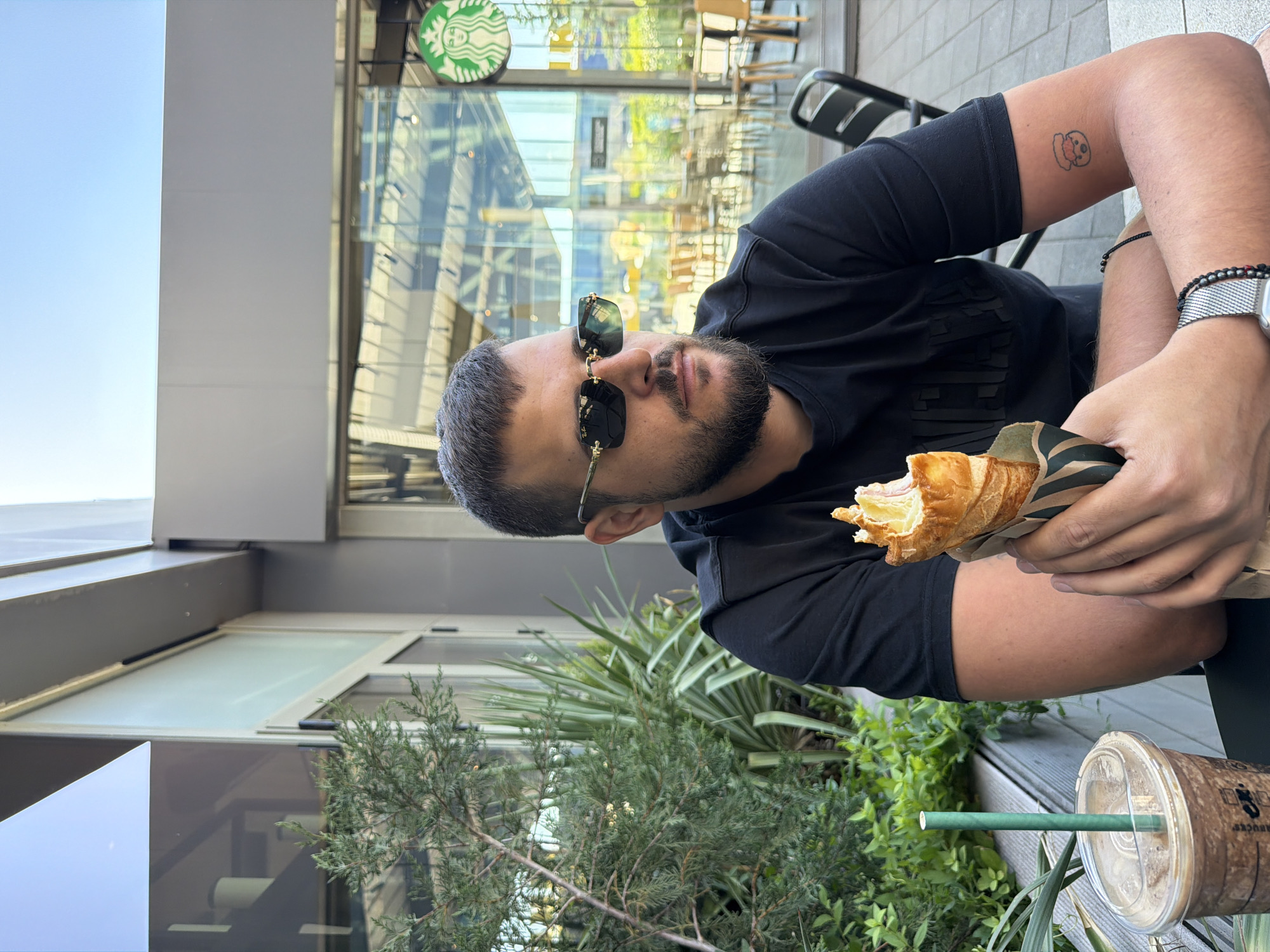 A man sits at a table outdoors, holding a partially eaten croissant in his left hand. He wears a black t-shirt and sunglasses. On his left arm, there is a tattoo. A Starbucks cup with a green straw is visible on the table next to him. A Starbucks store is visible in the background. The picture was taken in Sofia, Bulgaria.