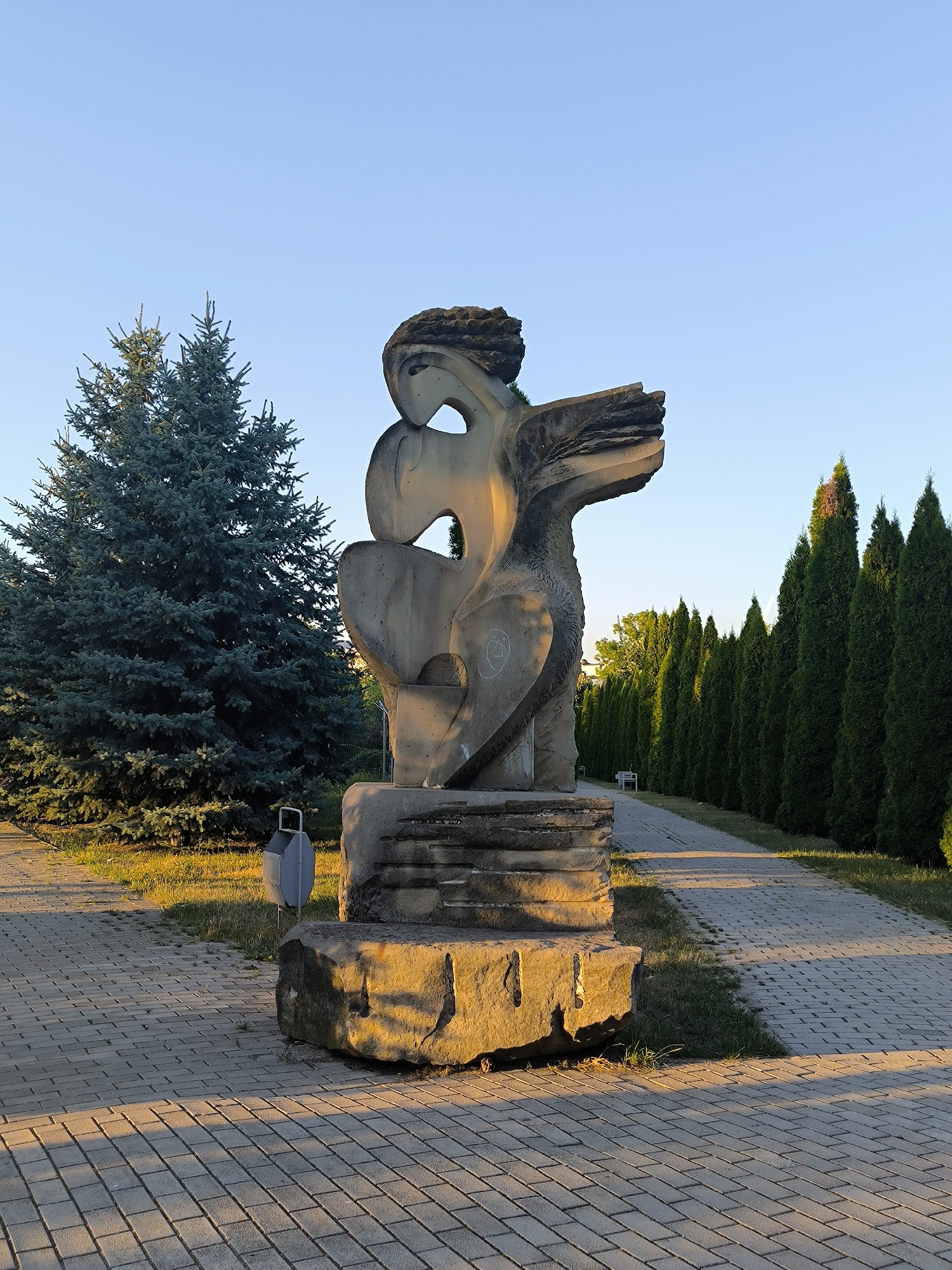 The image depicts an outdoor scene bathed in the golden light of a setting sun. In the foreground, a modern abstract sculpture made of stone stands on a paved walkway. The sculpture is characterized by its angular, fragmented forms, with a prominent void in the center and what appears to be wings or stylized plumage. The base of the sculpture rests on a large, rough-hewn rock.
To the left of the sculpture is a large, dark green conifer tree, possibly a spruce. Adjacent to the sculpture and slightly behind it, a modern, gray trash receptacle is visible. The walkway itself is constructed of interlocking gray pavers, and to the right, a neatly trimmed row of dark green cypress trees forms a dense hedge along a winding path. A park bench can be seen in the distance, nestled among the cypress trees. The background consists of a clear, pale blue sky.
This sculpture is located in the Park "La Izvor" in Chișinău, Moldova. It is attributed to the sculptor Ion Bolocan and dates from 2002, according to inscriptions on the sculpture. The overall atmosphere is peaceful and serene, highlighting the interplay of light and shadow on the artwork and its surroundings.