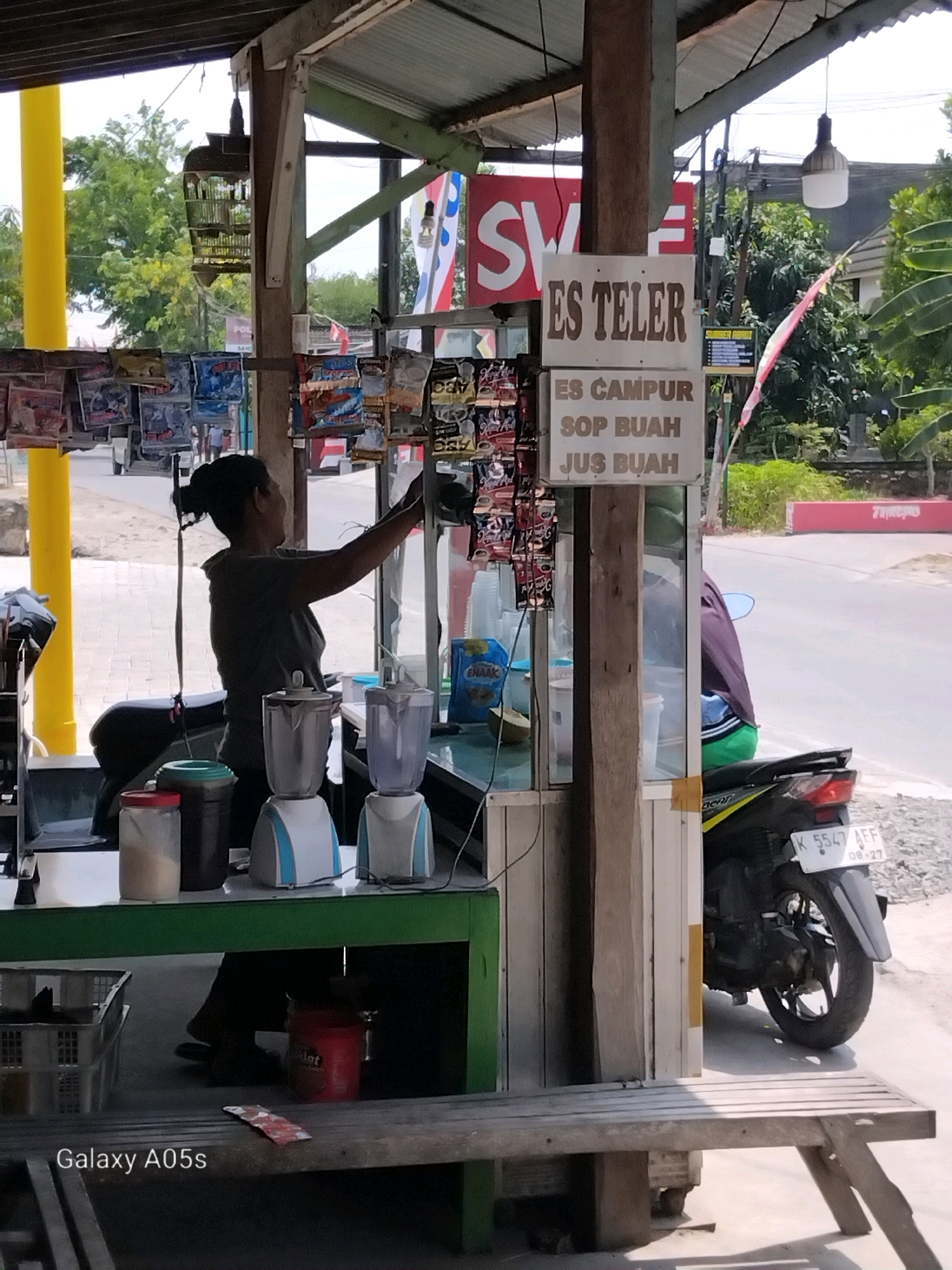 A street vendor with dark hair pulled back in a bun is standing behind a green counter in Karangayung, Indonesia. She is wearing a dark grey t-shirt. In front of her on the counter are two blenders, one white and blue, and another blue. To the left of the blenders are two containers, one clear with a red lid and a white powdery substance inside, and another dark container with a green lid. A motorcycle with a white license plate that reads "K 5547 AFF 08-27" is parked to the right of the vendor. Behind the vendor, a sign displays "ES TELOR" and "ES CAMPUR SOP BUAH JUS BUAH". The environment appears to be an outdoor market or roadside stall, with a corrugated metal roof overhead and a wooden bench in the foreground. Trees and buildings are visible in the background, suggesting a semi-urban or rural setting. The lighting indicates it is daytime, with sunlight casting shadows.