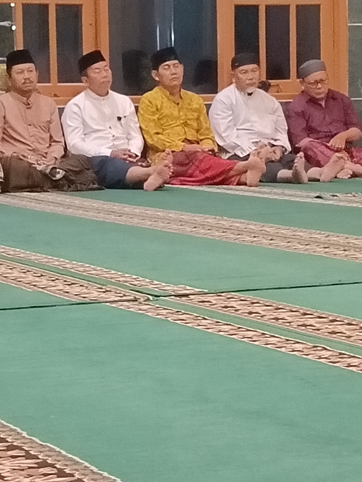 Six men are seated on a green carpet with a patterned border. The men are wearing traditional Islamic clothing, including peci hats. They appear to be in a place of worship, possibly a mosque, given the carpet and their attire. The men are facing towards the viewer, though their expressions are not clearly discernible. The lighting suggests an indoor setting, possibly at night or in a dimly lit room. The background shows wooden paneled windows. The image is taken from a low angle, looking up at the men.