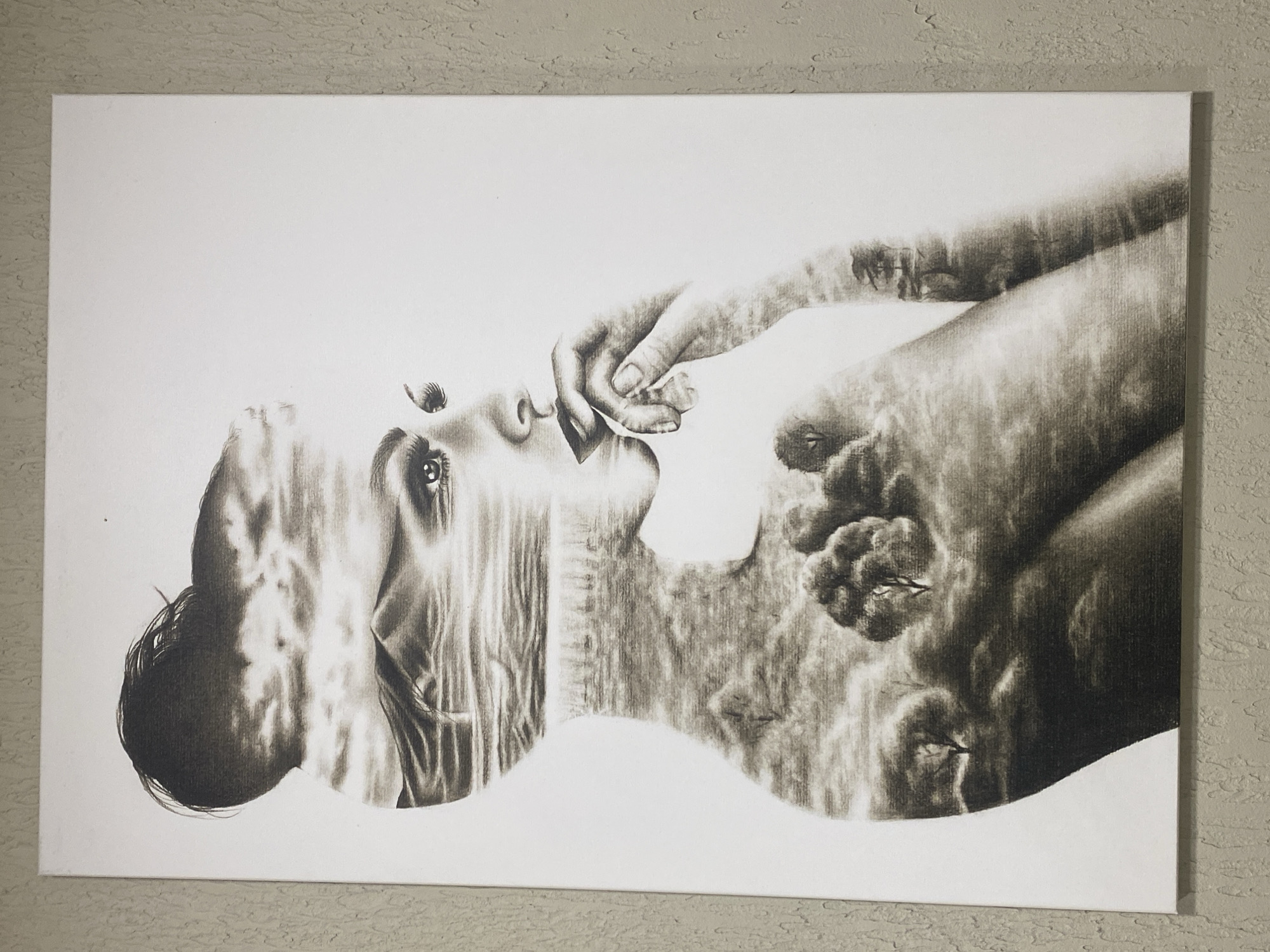This is a charcoal drawing on canvas featuring a profile view of a woman's head and upper body. The drawing is a double-exposure style, with a landscape scene integrated into the silhouette of the woman. The landscape includes elements like clouds, mountains, trees, and what appears to be a body of water. The woman's facial features, such as her eyes, nose, and mouth, are visible and rendered with detail. Her hand is raised to her mouth, suggesting contemplation or a pensive mood. The background of the canvas is white, and the drawing is framed by a textured wall, possibly a stucco finish, which appears to be a neutral gray or beige color. The lighting in the image highlights the drawing and the textured wall. There is no indication or identifiable elements within the image that would suggest a specific location in Kemer, Turkey.