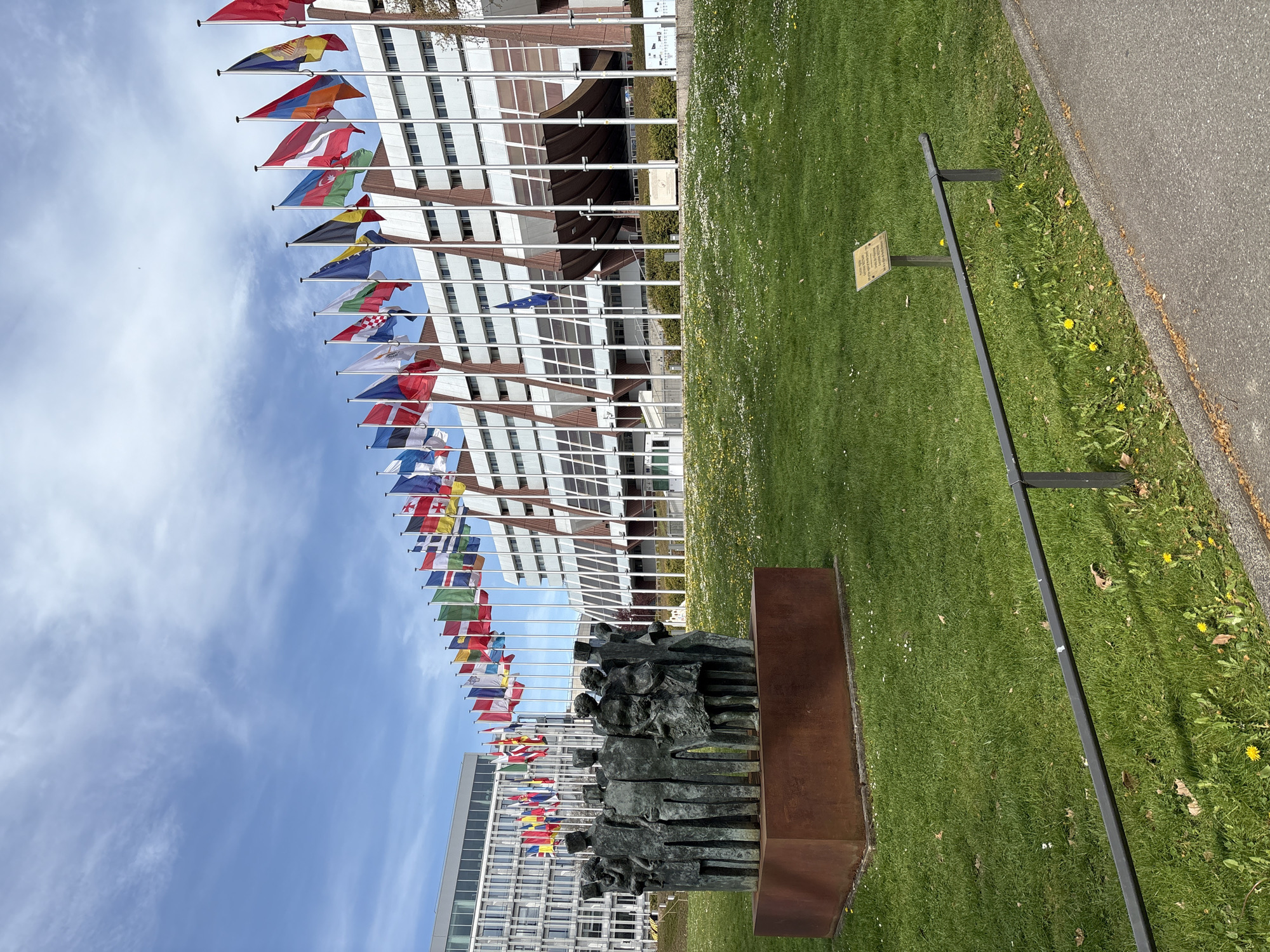 The image shows a view in Strasbourg, France, featuring a building with numerous white-framed windows. A row of flagpoles displays various national flags, creating a colorful display against the backdrop of the building and a blue sky with some clouds. In the foreground, there is a sculpture of a group of people rendered in a dark, possibly bronze, material. The sculpture is mounted on a dark base. A grassy lawn is to the right of the sculpture. Along the right edge of the grass, there is a metal railing and a sidewalk. A small sign or plaque is also visible on the lawn near the railing.