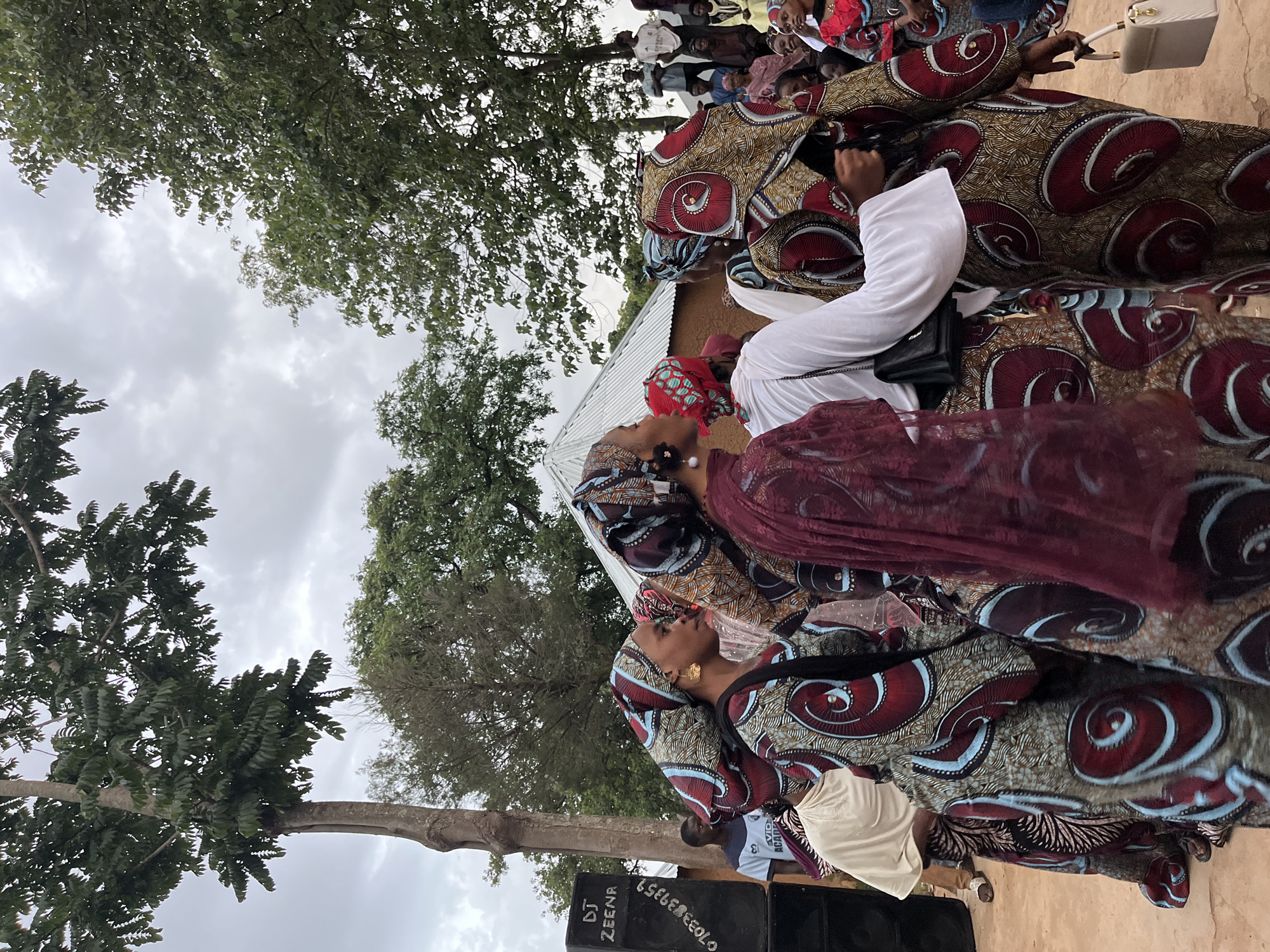 The image depicts an outdoor gathering, possibly a celebration, in Jos, Nigeria. Several individuals are the central focus, predominantly women adorned in vibrantly patterned traditional attire. The clothing features intricate designs in colors like maroon, blue, gold, and brown, with headscarves matching or complementing their outfits. One woman in the foreground wears large gold hoop earrings.
To the left, a large black speaker system with "DJ ZEENR" and a phone number visible on its side suggests a potential DJ setup for the event. Behind the speaker, a man in a blue t-shirt is visible. In the background, there's a glimpse of a building with a corrugated metal roof and other people gathered, some seated. Trees with lush green foliage frame the scene, and the sky is overcast with clouds. The ground appears to be dirt or sand. The atmosphere suggests a lively community event in an outdoor setting in Jos.