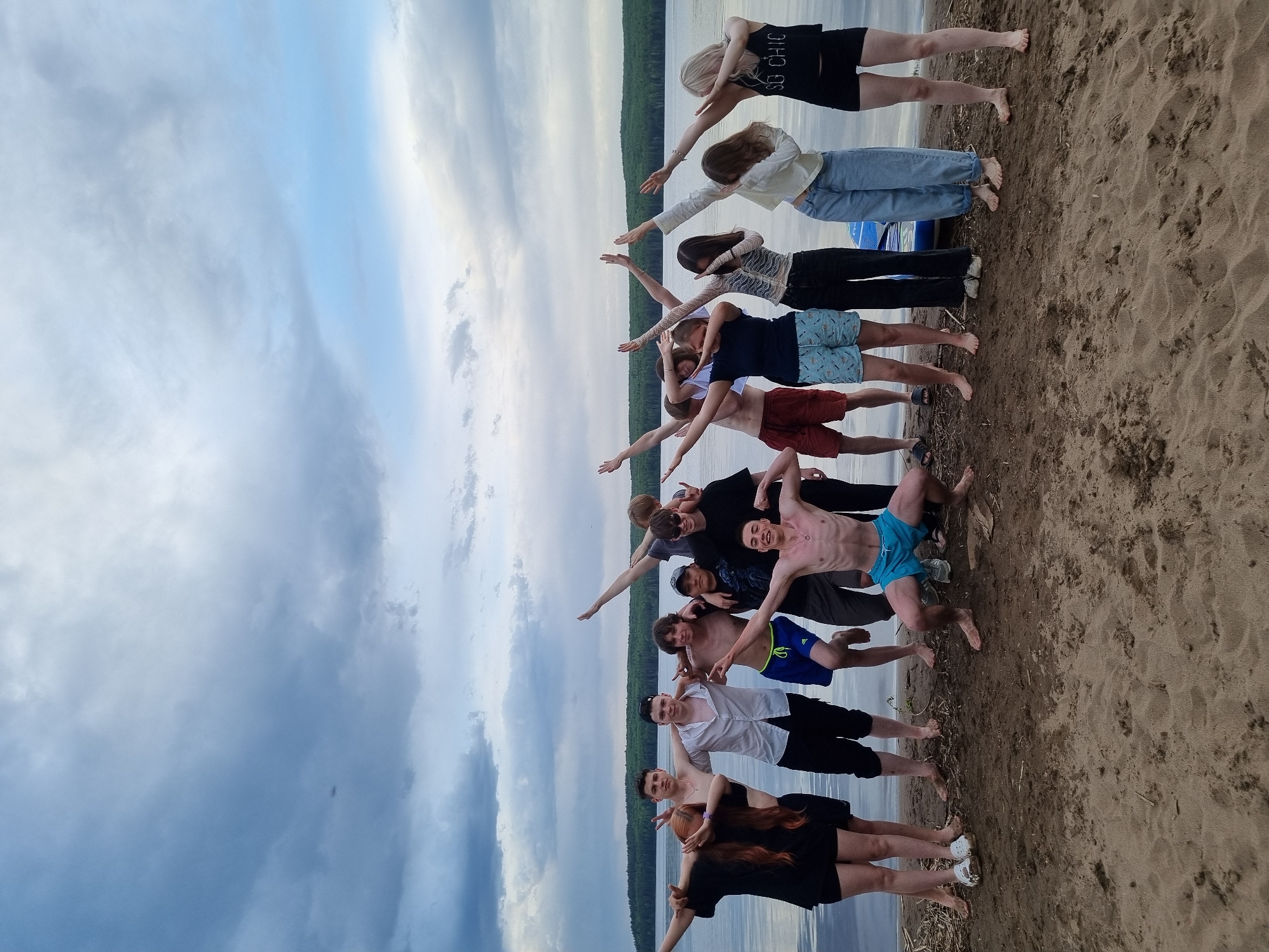 A group of approximately 15 people is lined up on a sandy beach, posing in a line with their arms extended as if forming a chain. The background features a body of water and a treeline. The location is Chaikovsky, Russia.